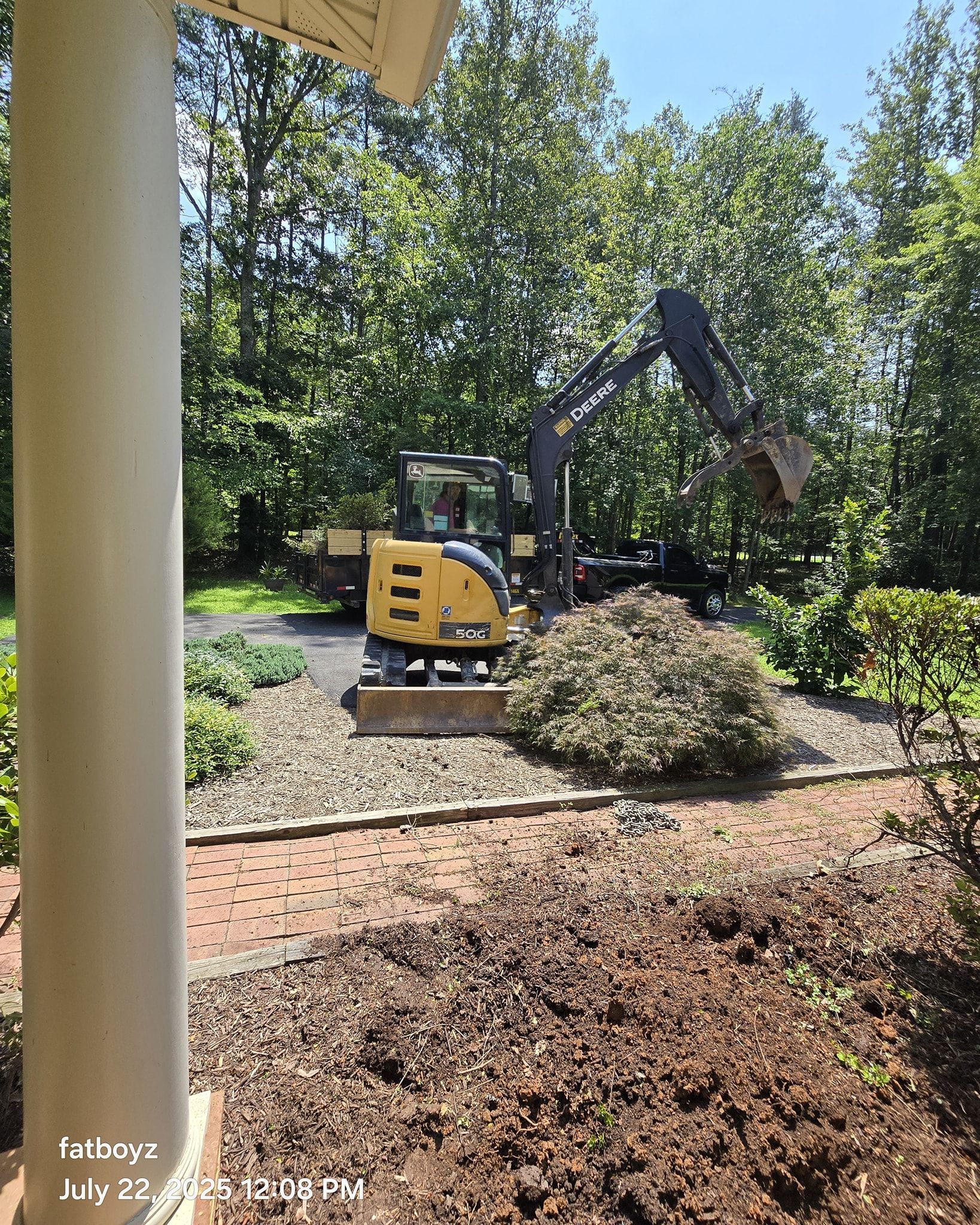 Small yellow excavator digging near shrubs on a landscaped path in a sunny park.