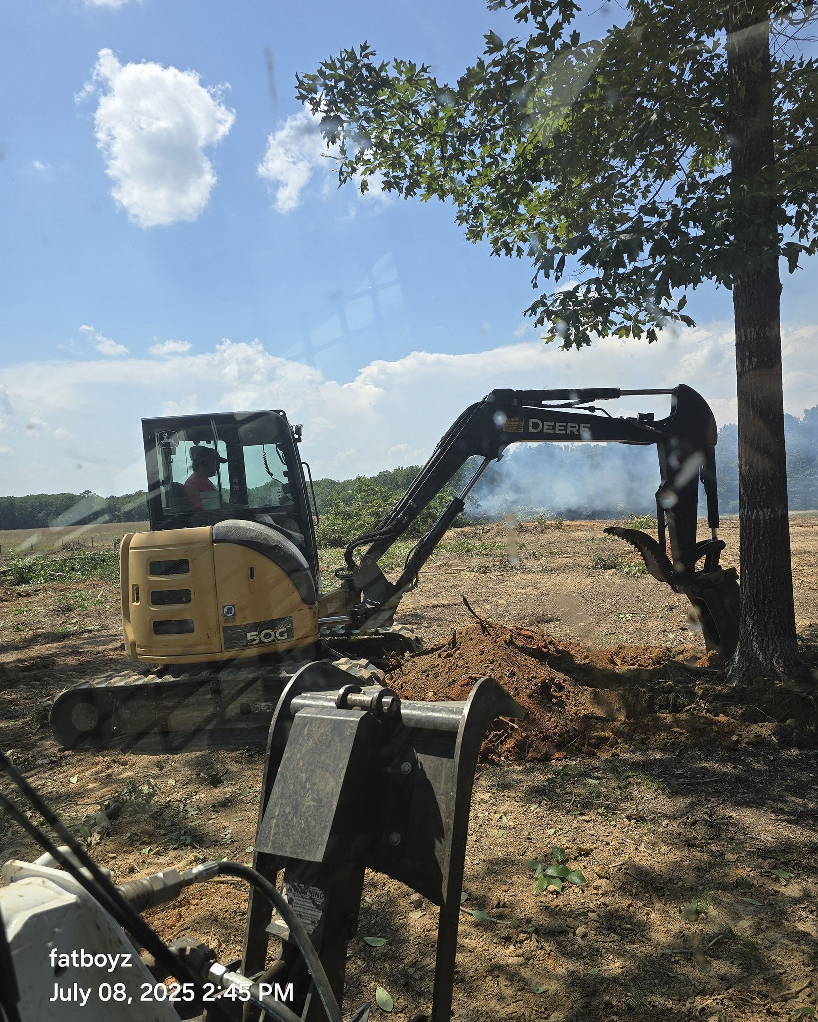 Yellow excavator digging soil in a field under a tree on a sunny day