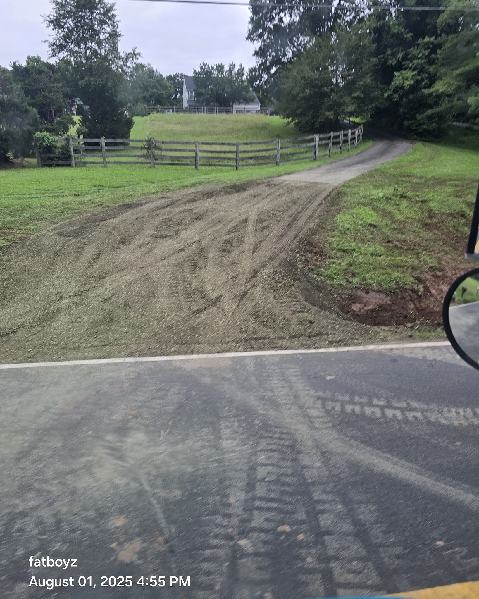 Dirt driveway leading to a wooden gate and grassy field, viewed from a paved road on a cloudy day