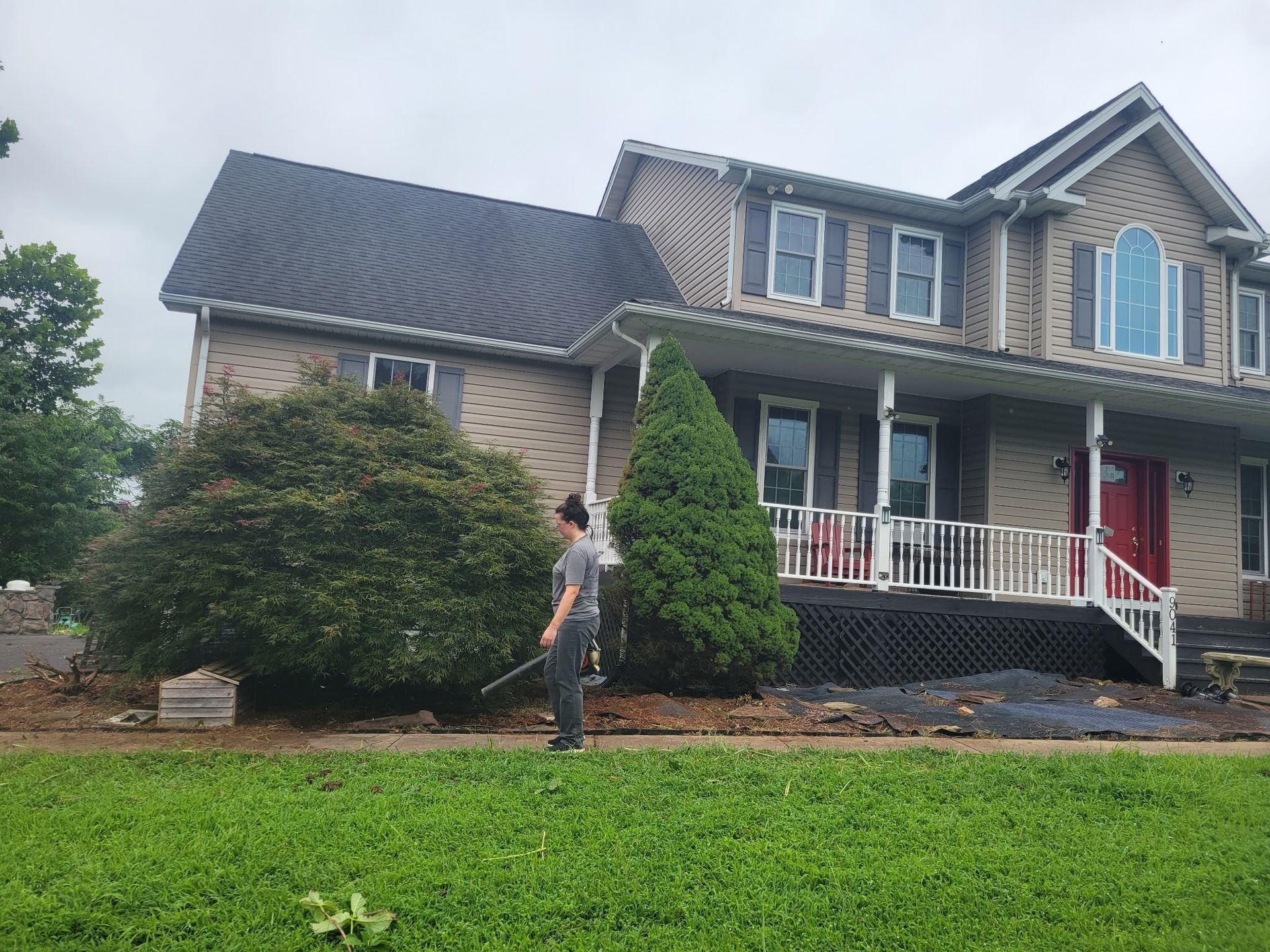 Person standing in front of a two-story gray house with a red front door and green lawn
