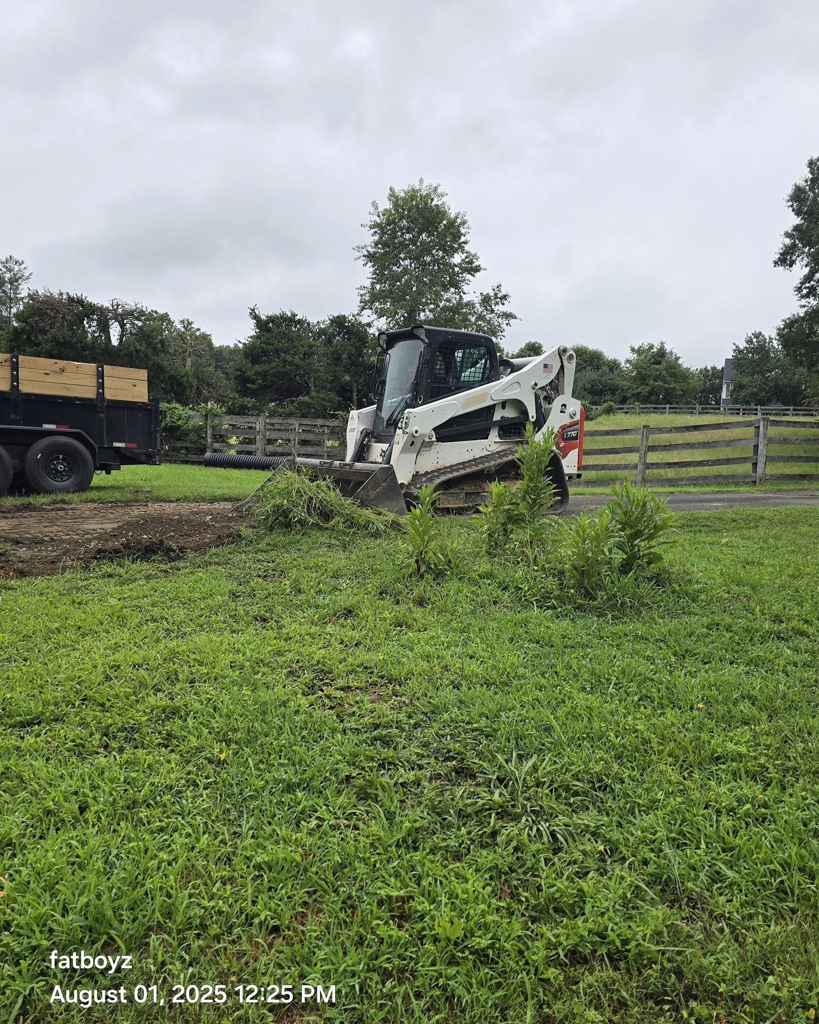 White skid-steer loader on a muddy field near a trailer, with a wooden fence and cloudy sky in the background.