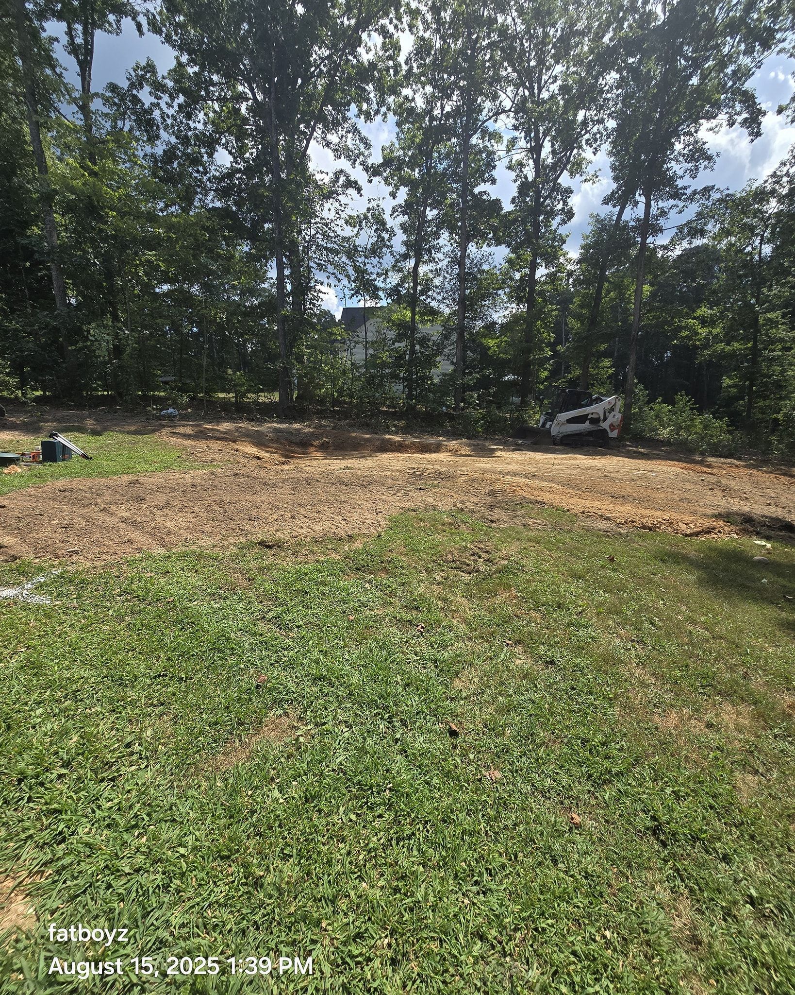 Grassy yard with a bare dirt patch under trees and scattered rocks near the edge.