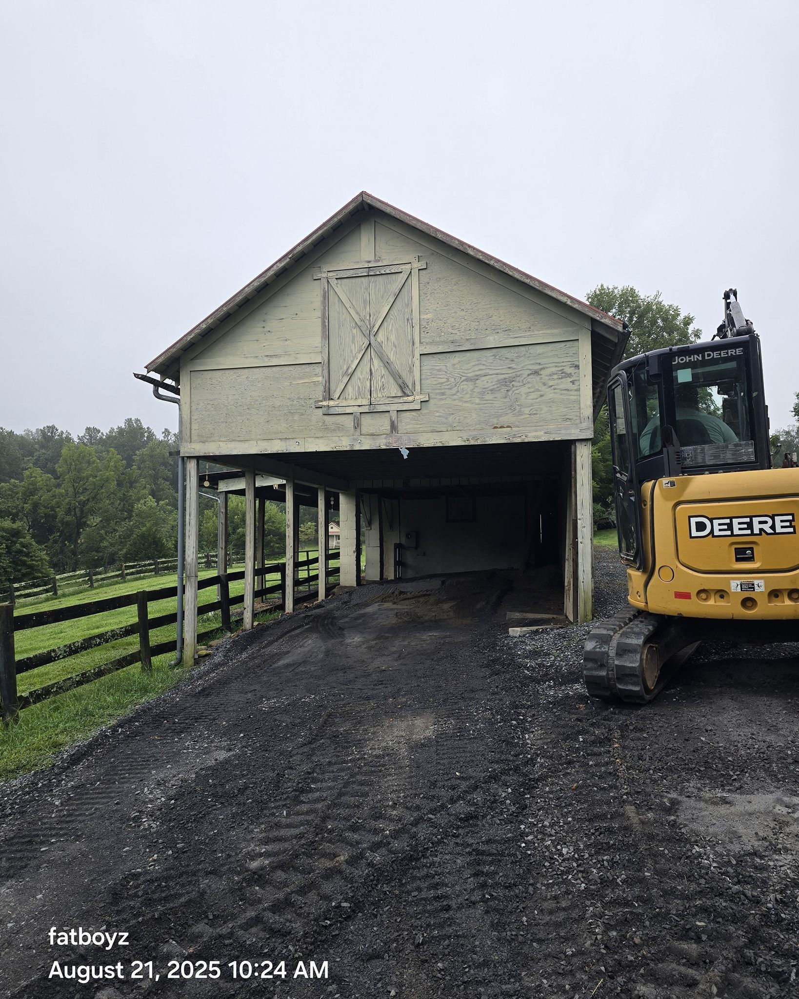 Yellow excavator beside an old barn on a gravel driveway in a rural wooded area