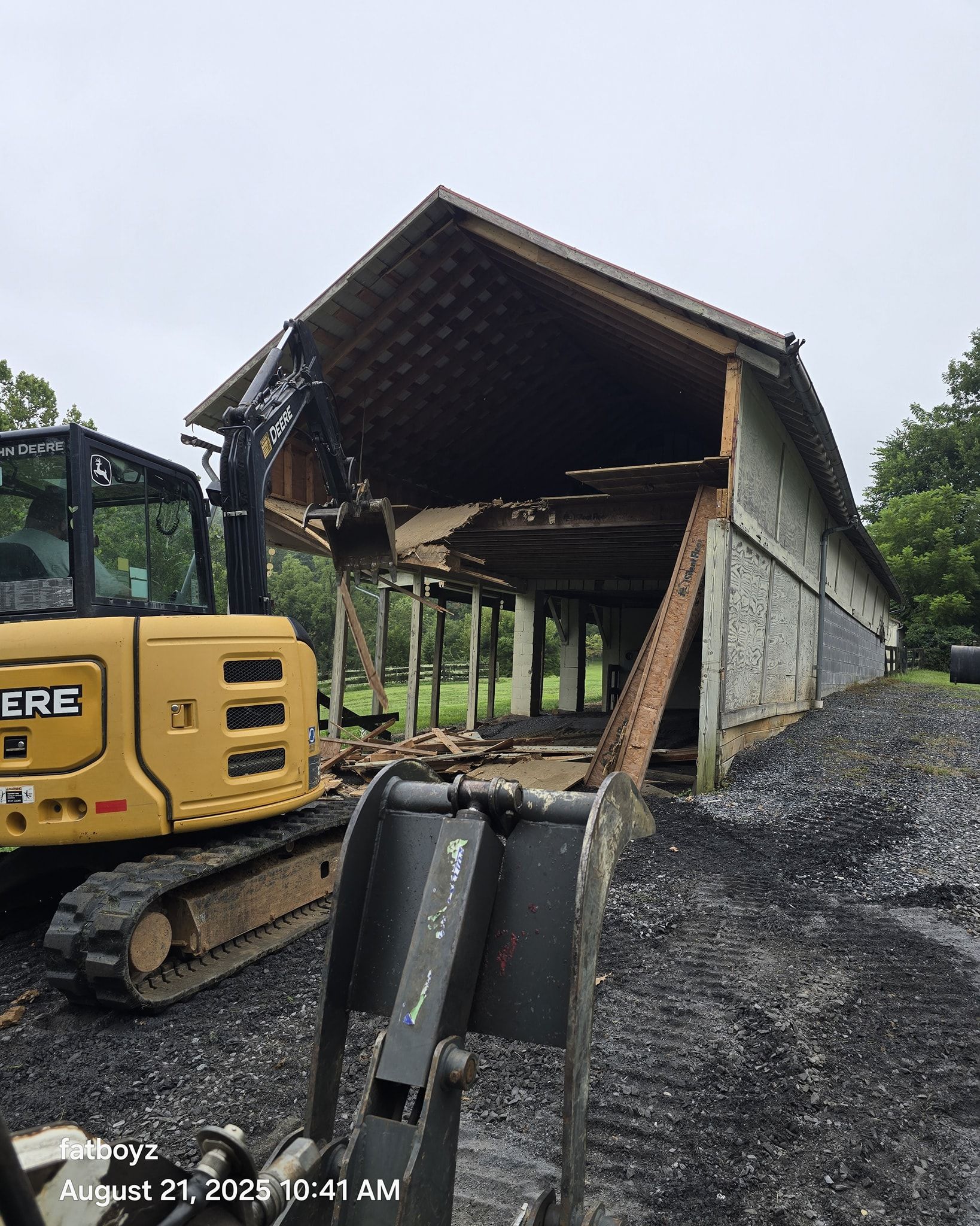 Yellow excavator beside a collapsed barn roof, with debris and gravel at a rural worksite