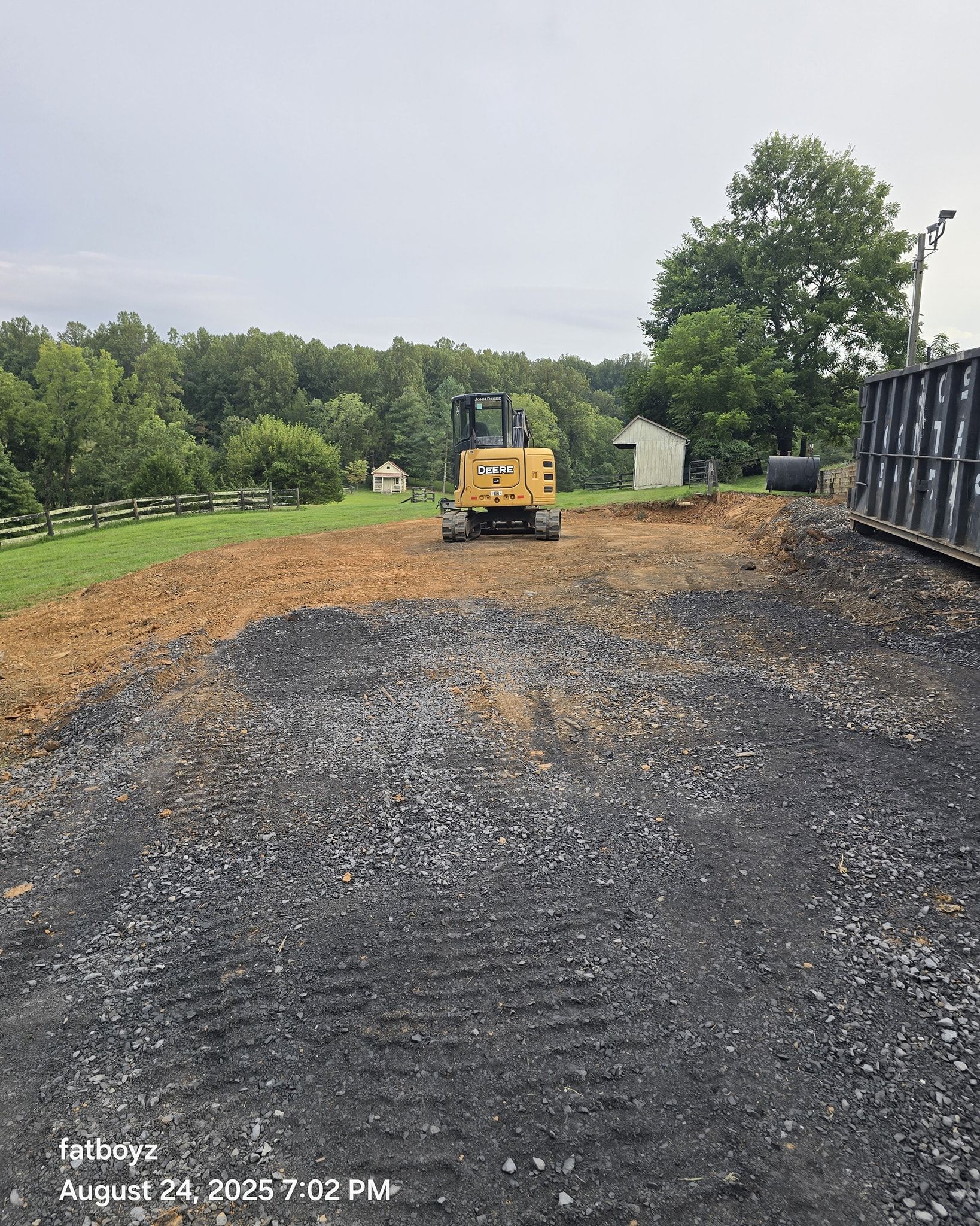 Gravel driveway under construction with a yellow loader, trees, and a white house in the background.