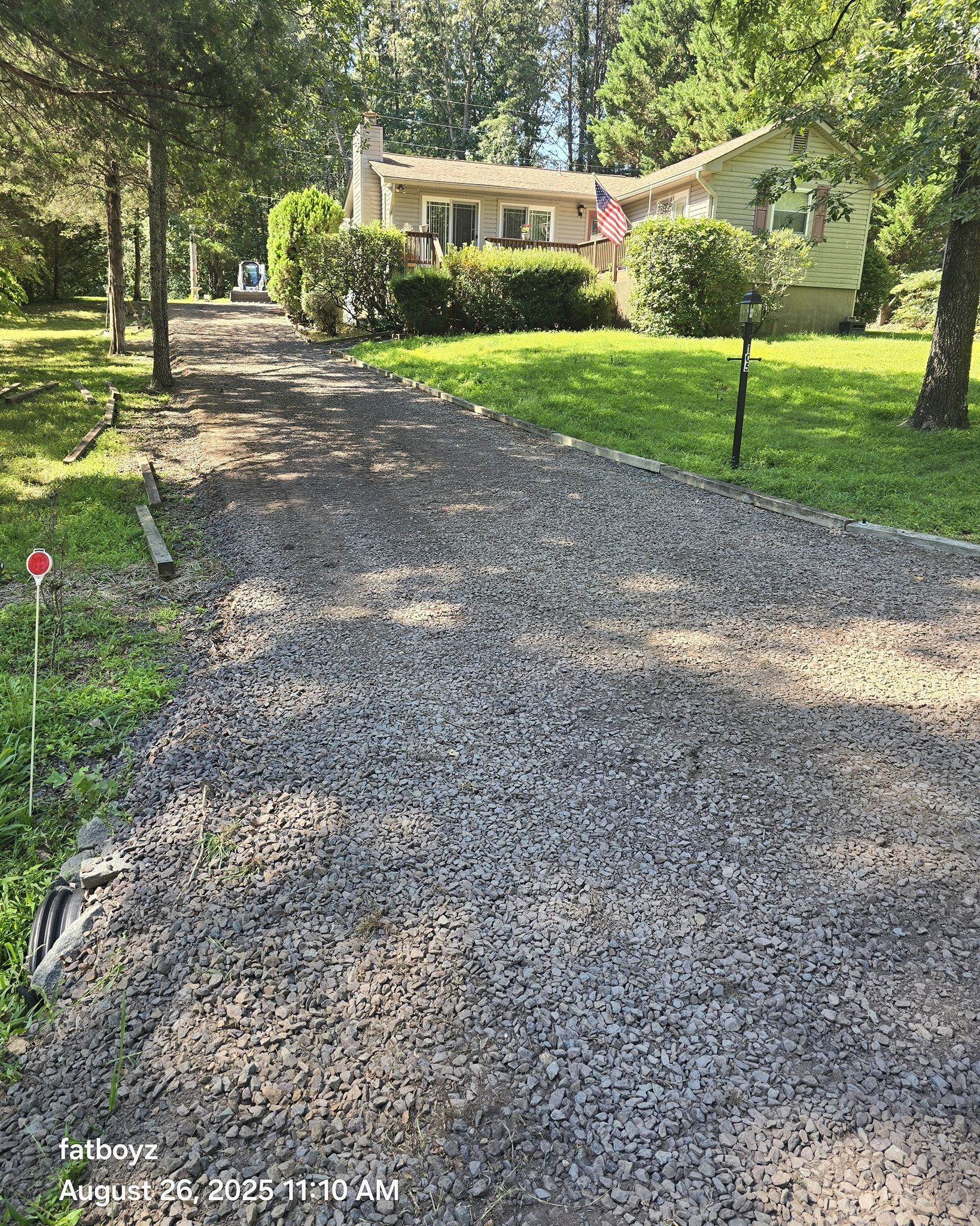 Gravel driveway leading to a beige house surrounded by tall green trees and lawn.