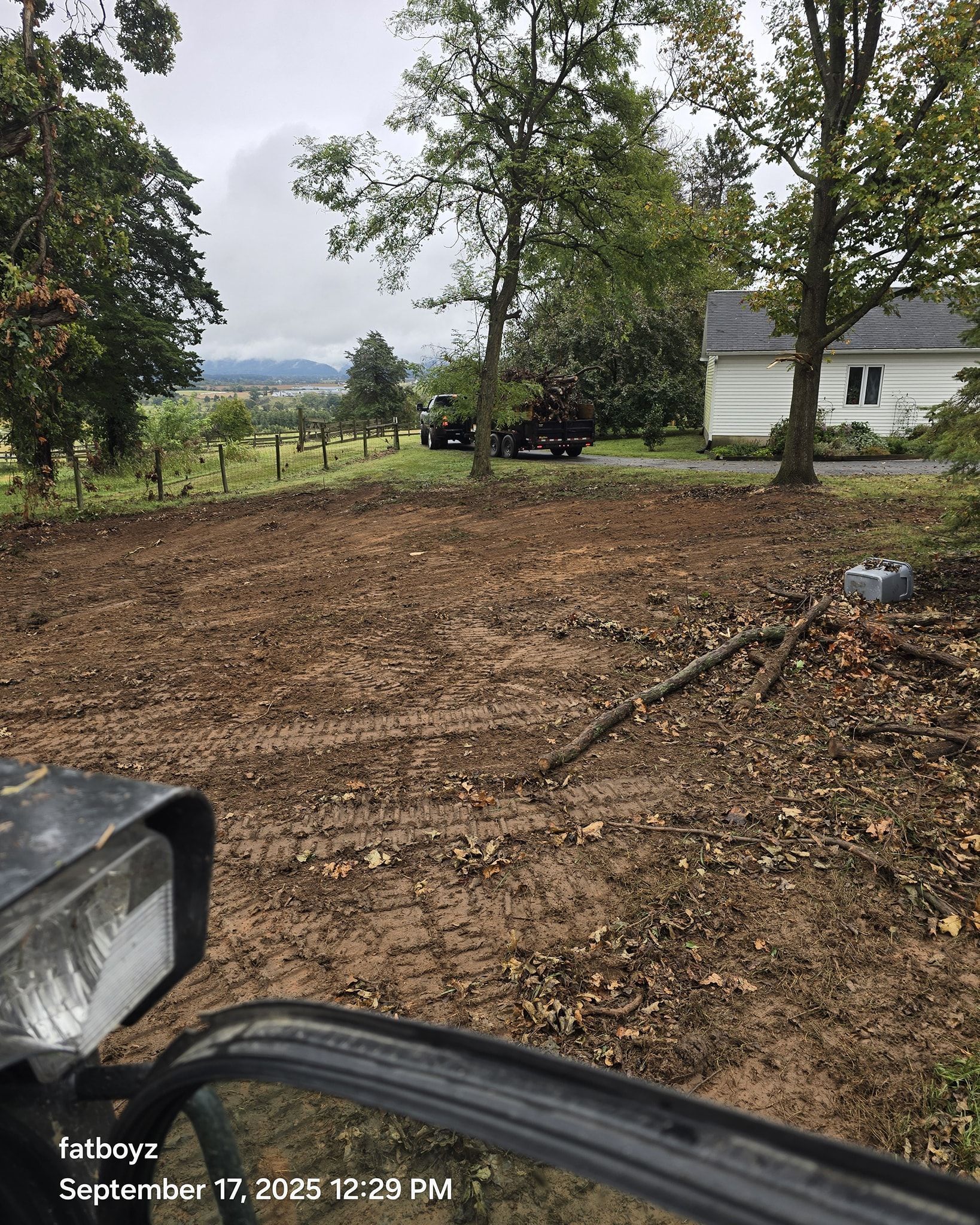 Dirt yard with scattered leaves, trees, and a white house in the background, viewed from a vehicle.