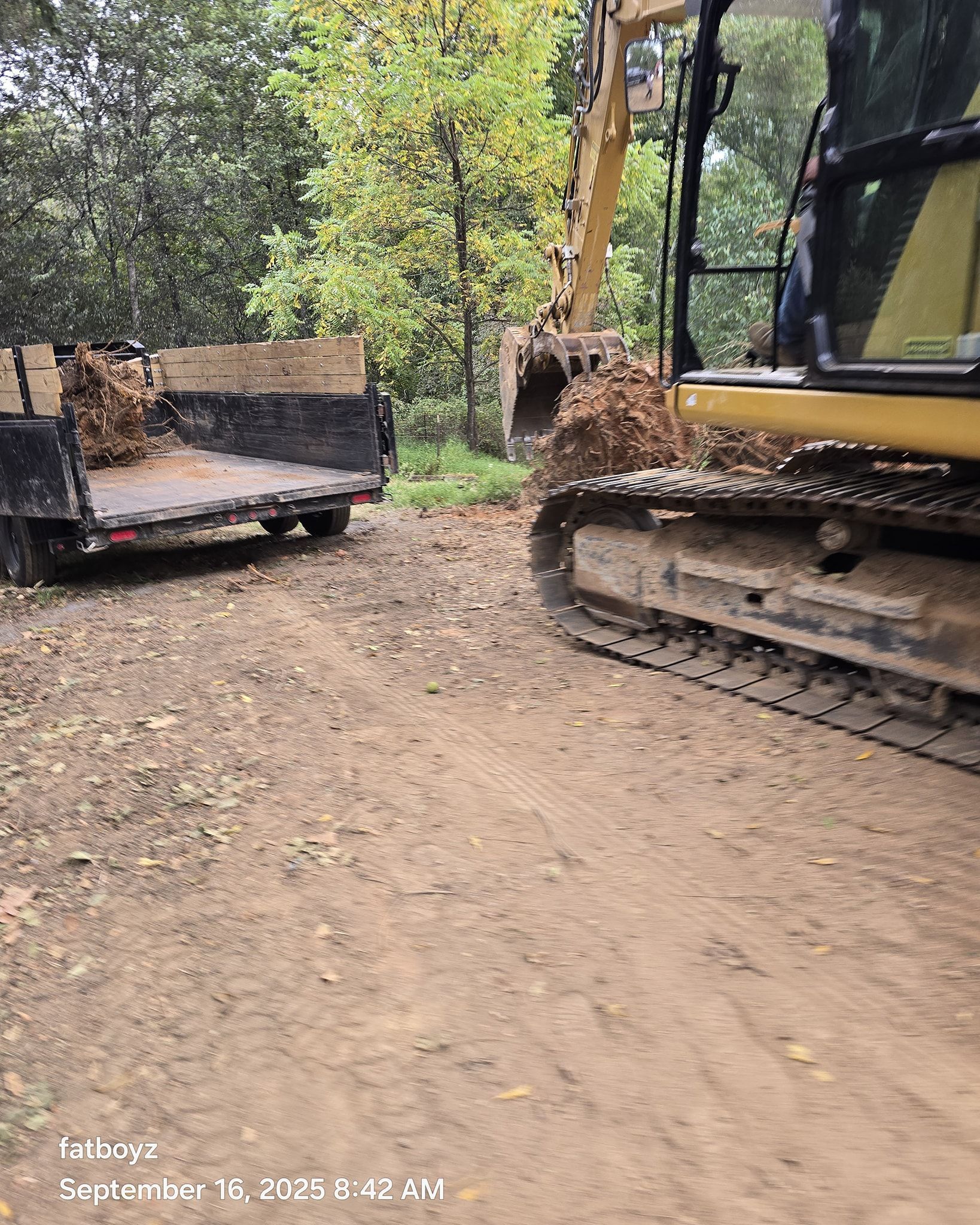 Yellow excavator on muddy forest road beside a flatbed truck and log pile.