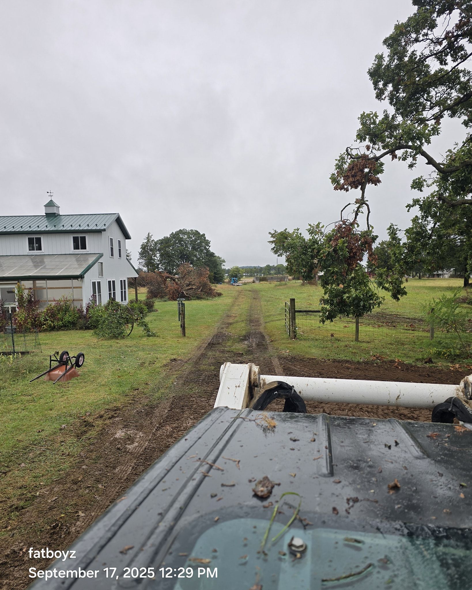 Rural dirt track beside a white house and trees under a cloudy sky, viewed from a vehicle