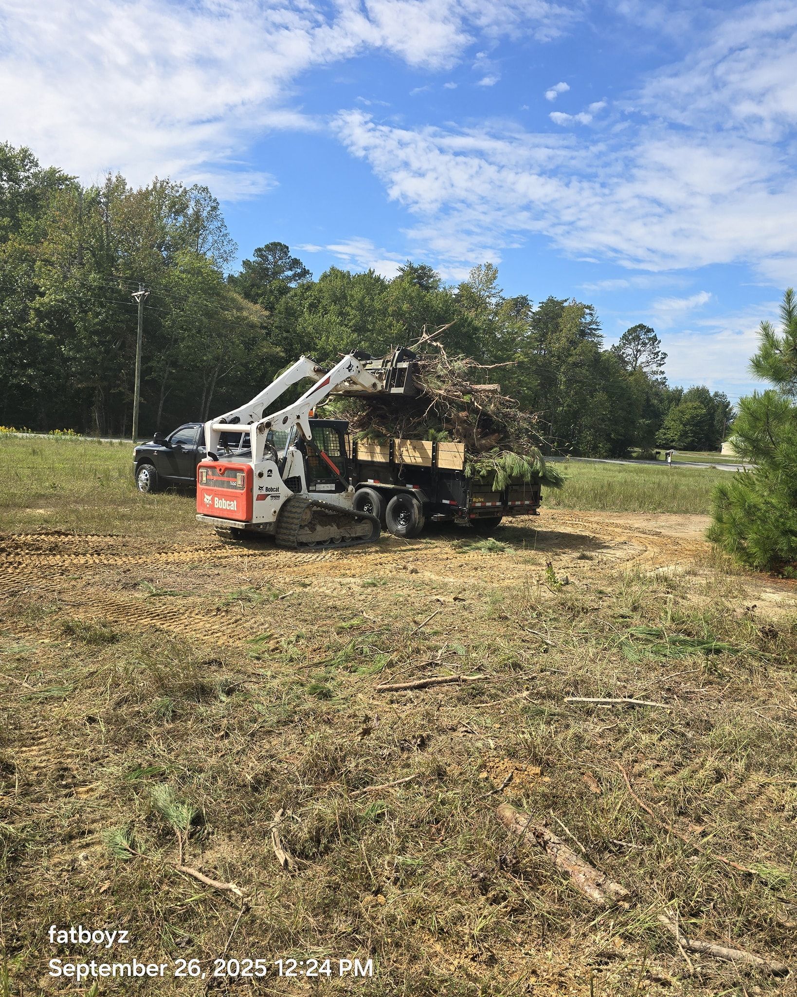 Orange excavator clearing cut brush in a grassy field with trees and blue sky in the background