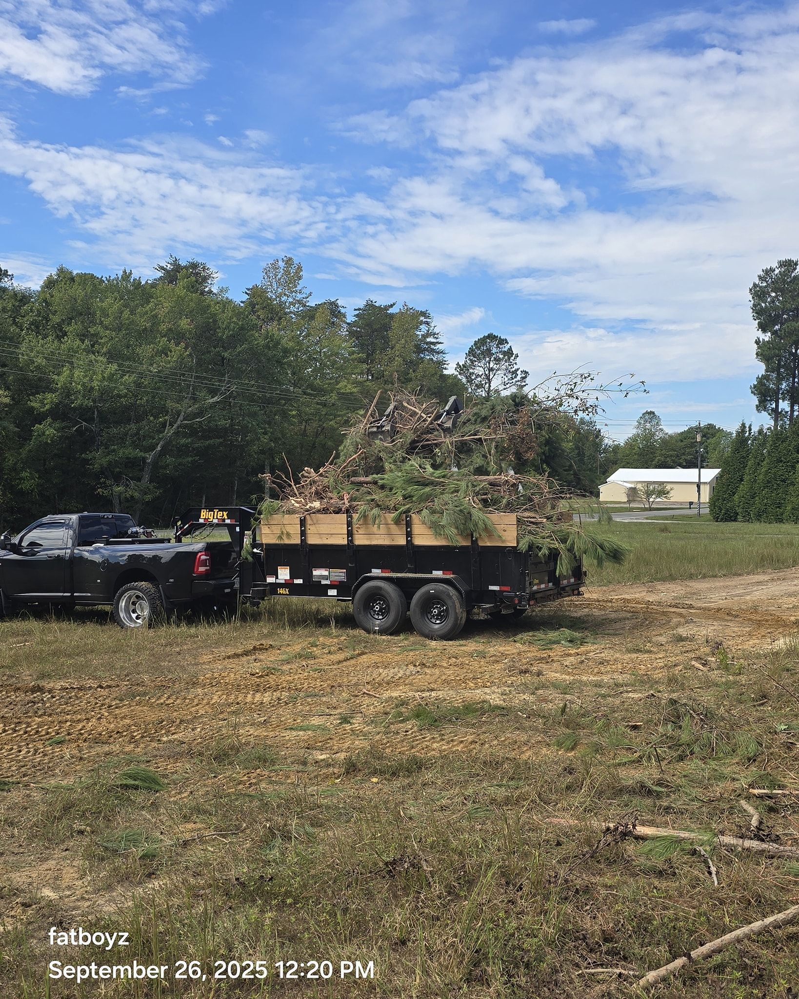 Black pickup towing a flatbed trailer loaded with brush in a sunny field near trees and a house.