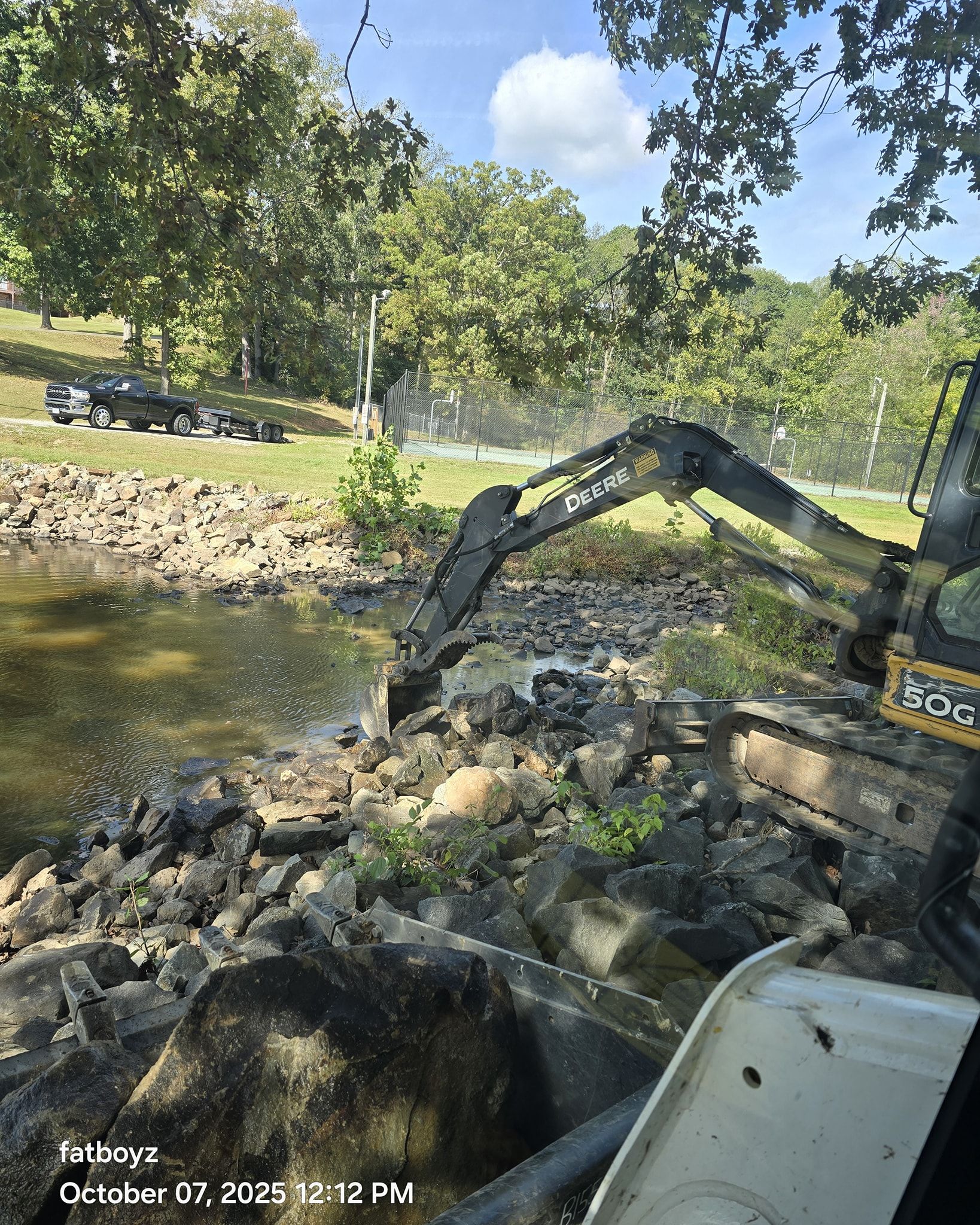 Excavator arm working in a rocky stream beside a tree-lined road on a sunny day
