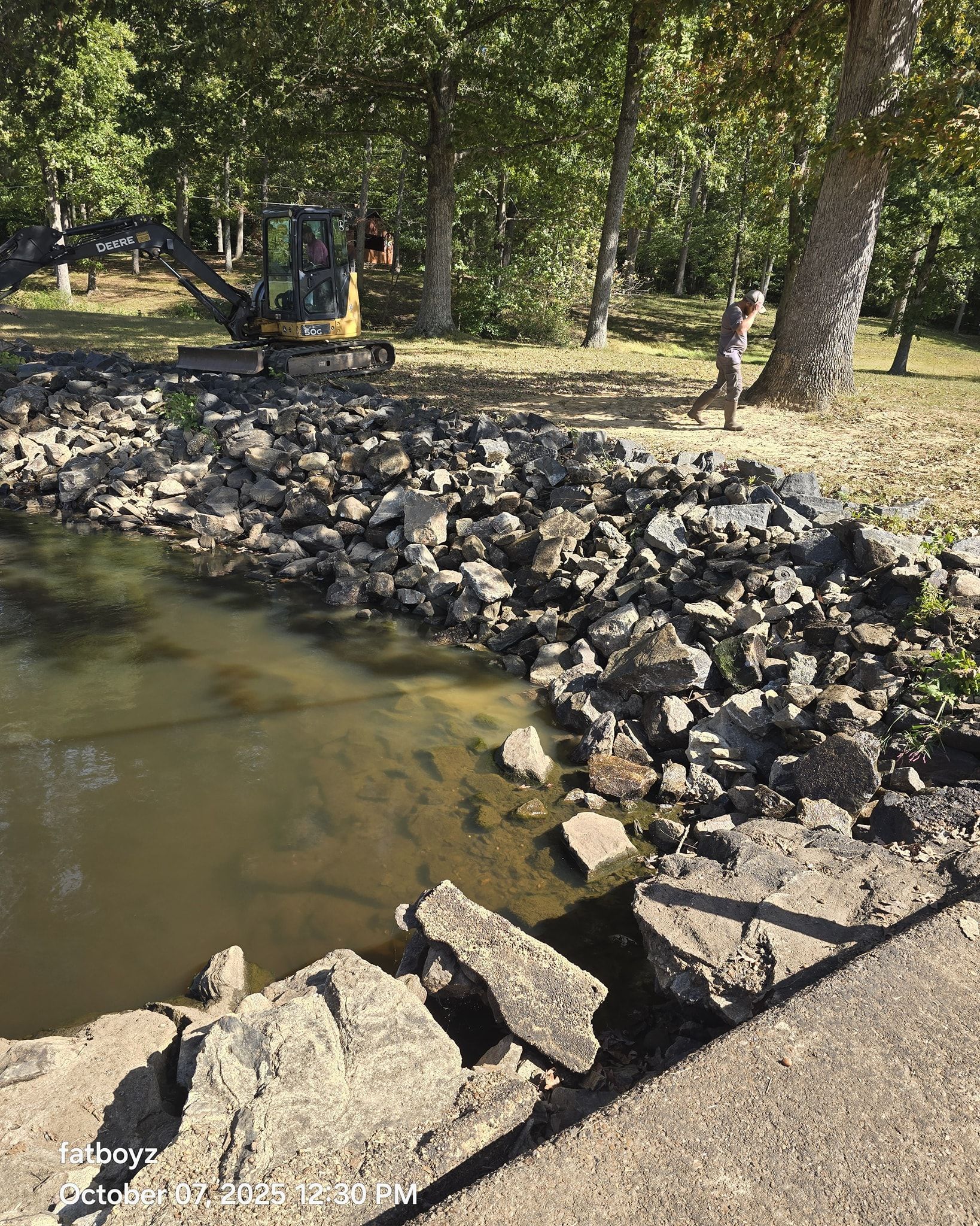 Rock-lined pond edge beside trees and a gravel path in a park