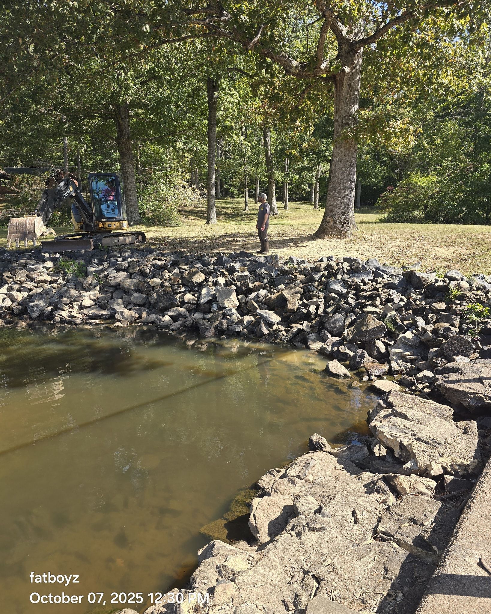 Rocky pond edge in a wooded park with shallow water and trees in the background.