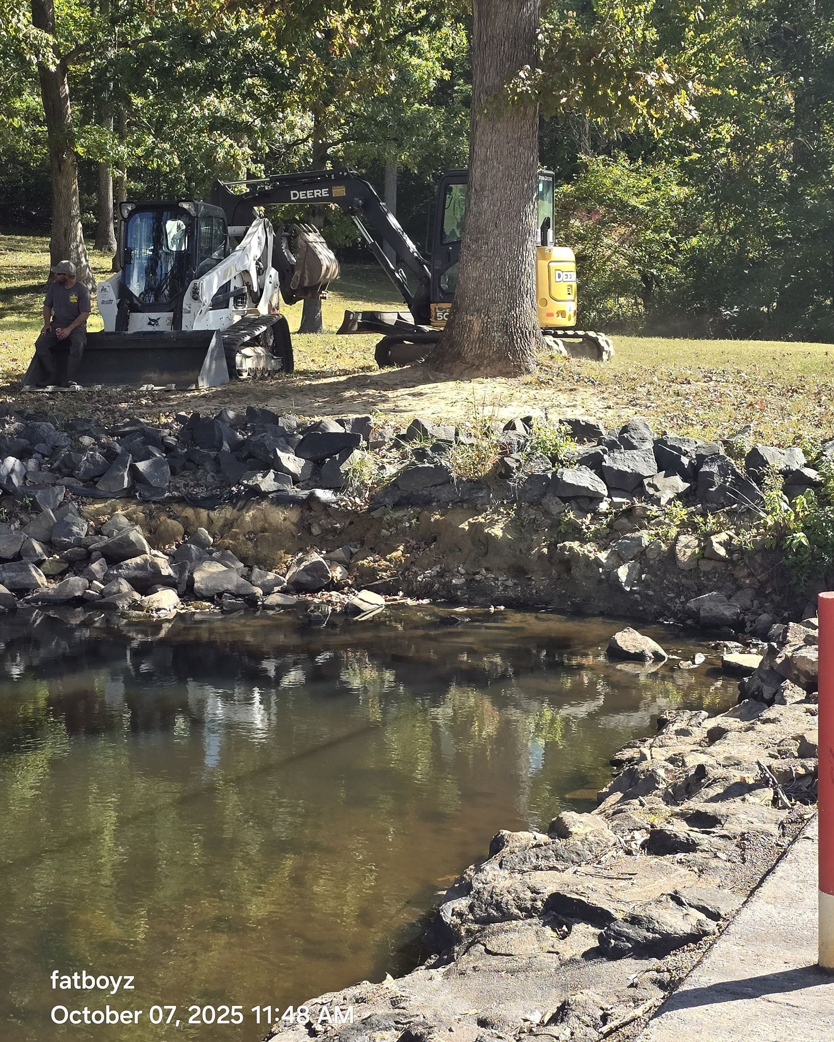 Rocky pond edge with calm water, trees, and a small excavator in the background.