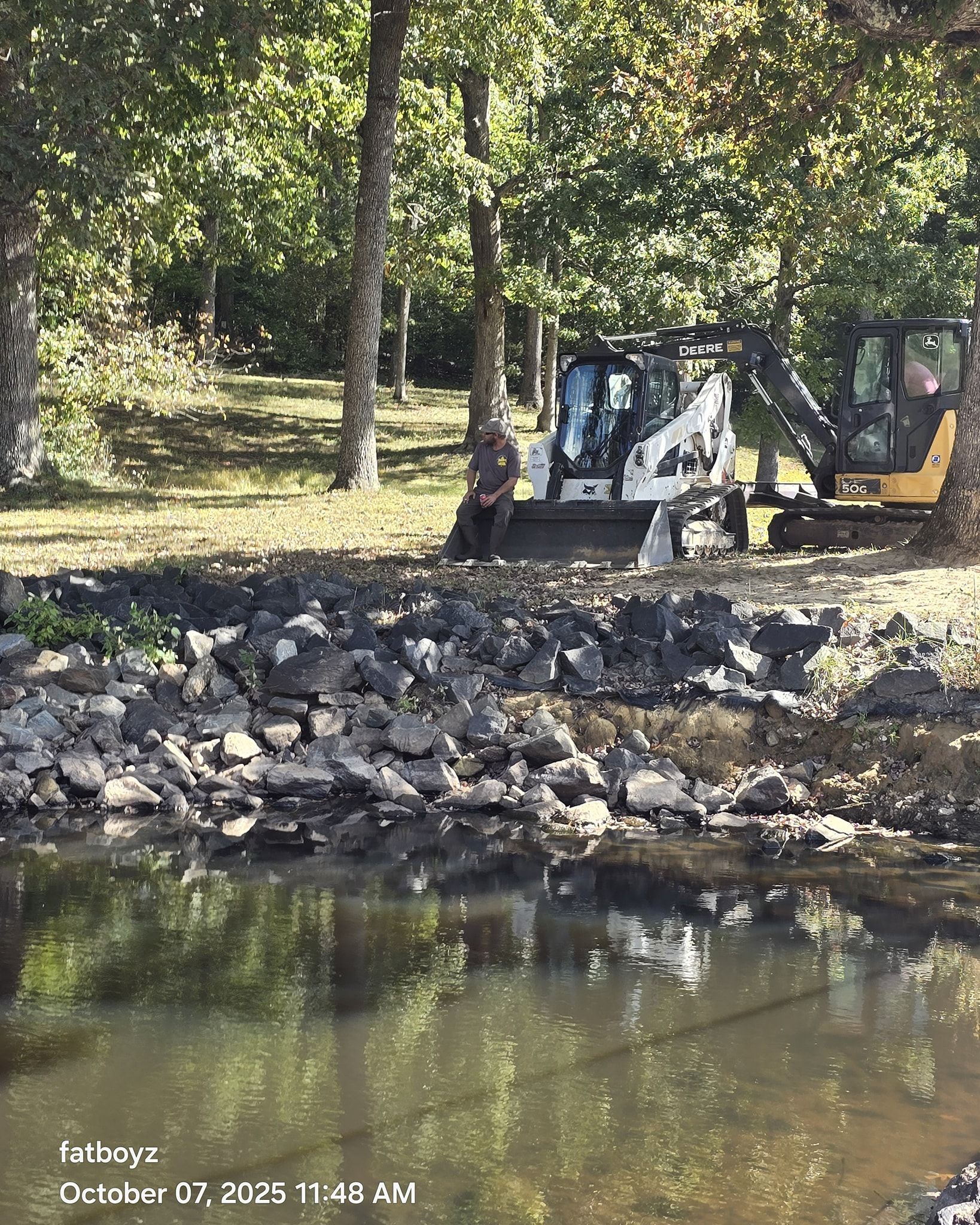 Small excavator and worker by a rocky riverbank in a wooded area.