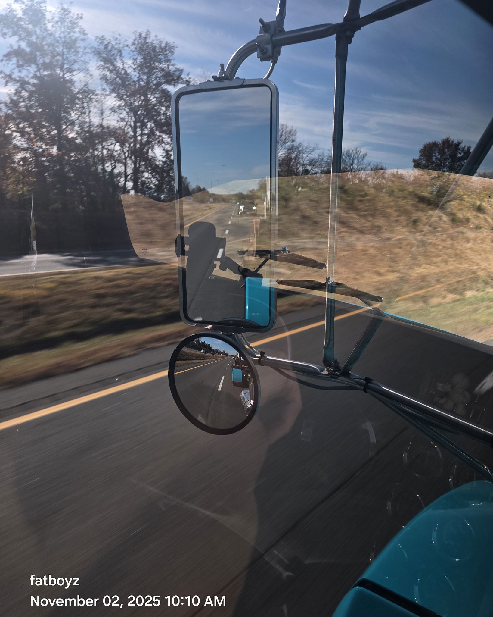 Truck mirror reflecting a road and roadside hillside, with a blue bottle in the cab and another vehicle visible.