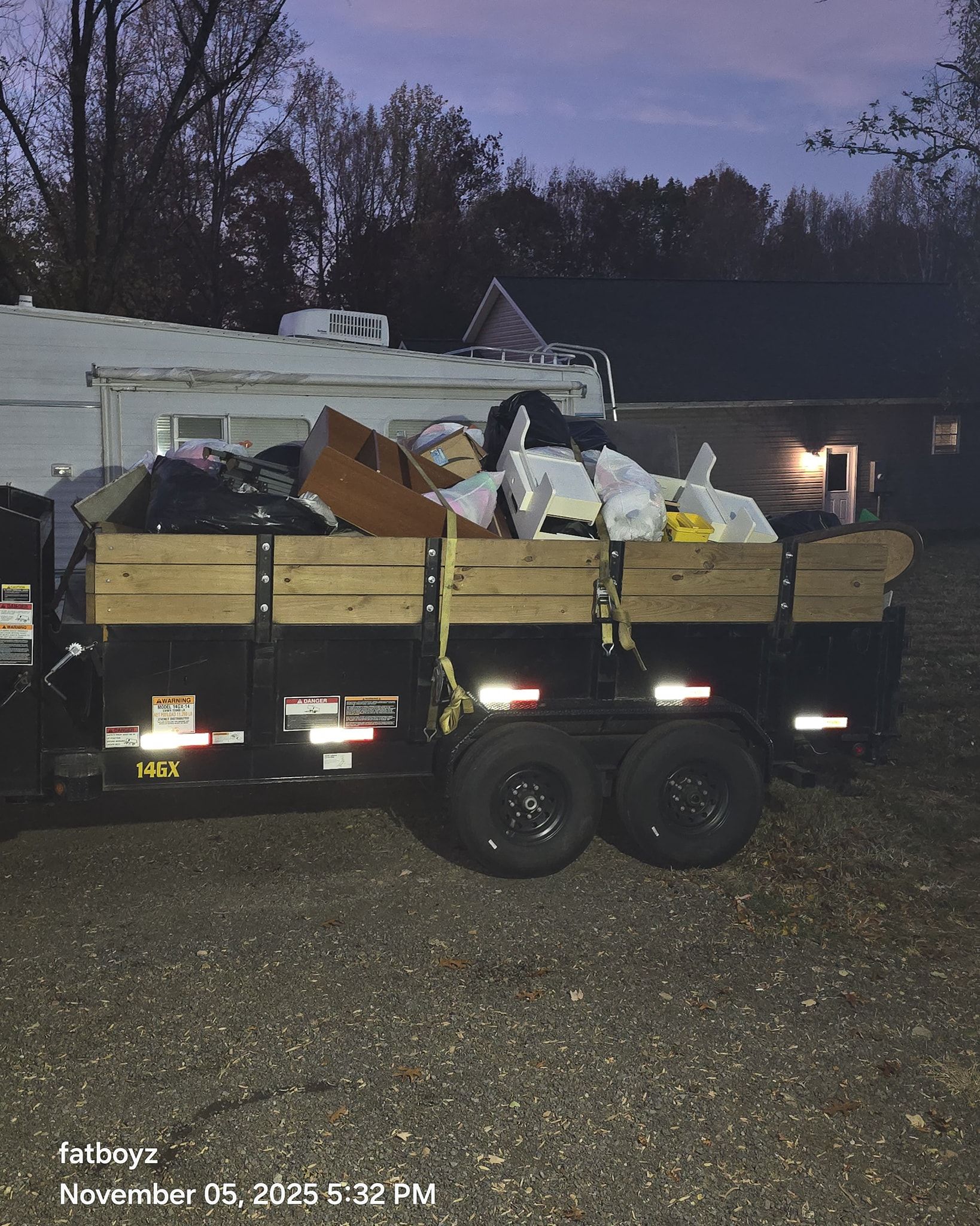 Loaded trailer with debris and household items parked on gravel at dusk near buildings.