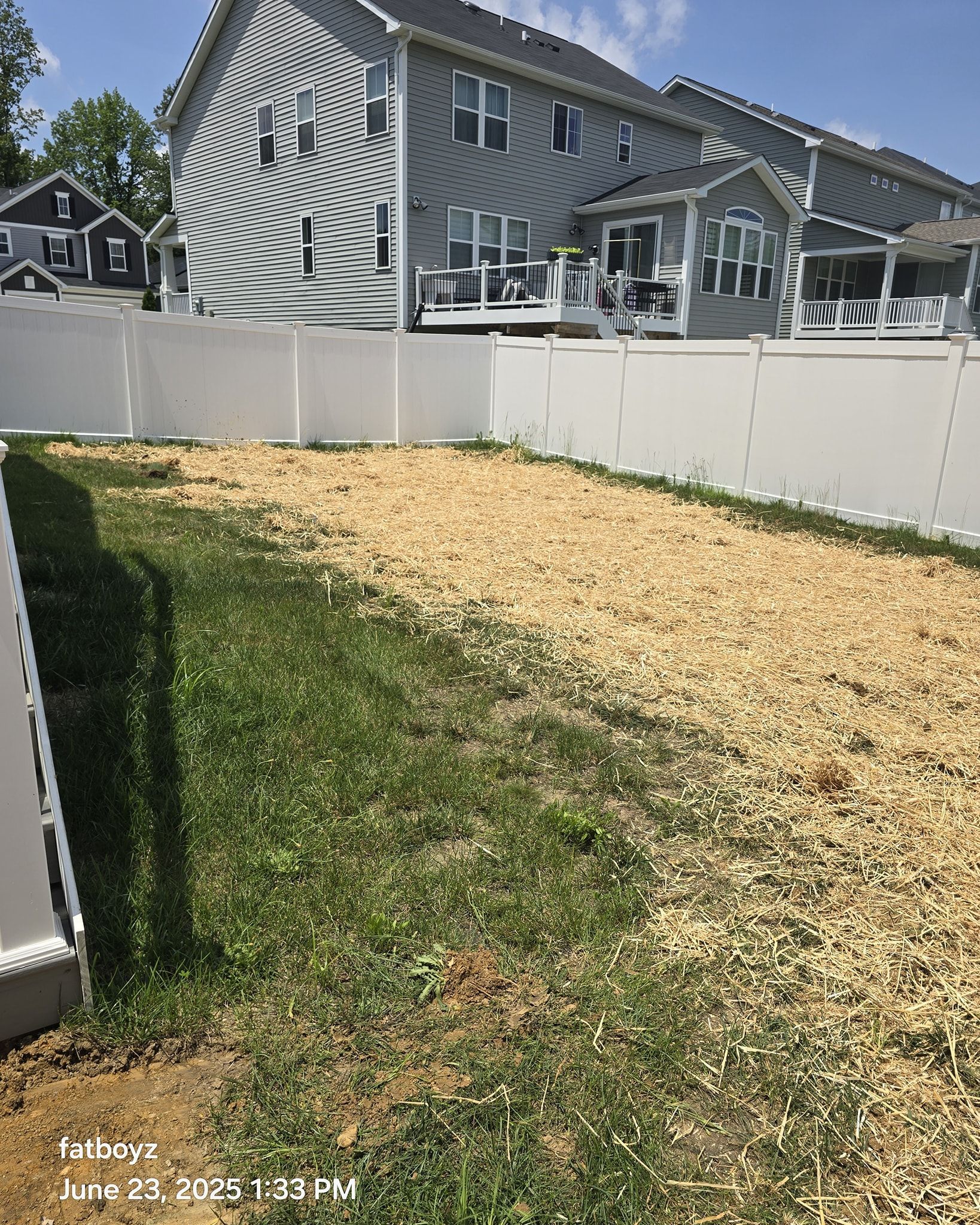 Backyard with white fence, green grass, and a large yellow straw-covered area under construction