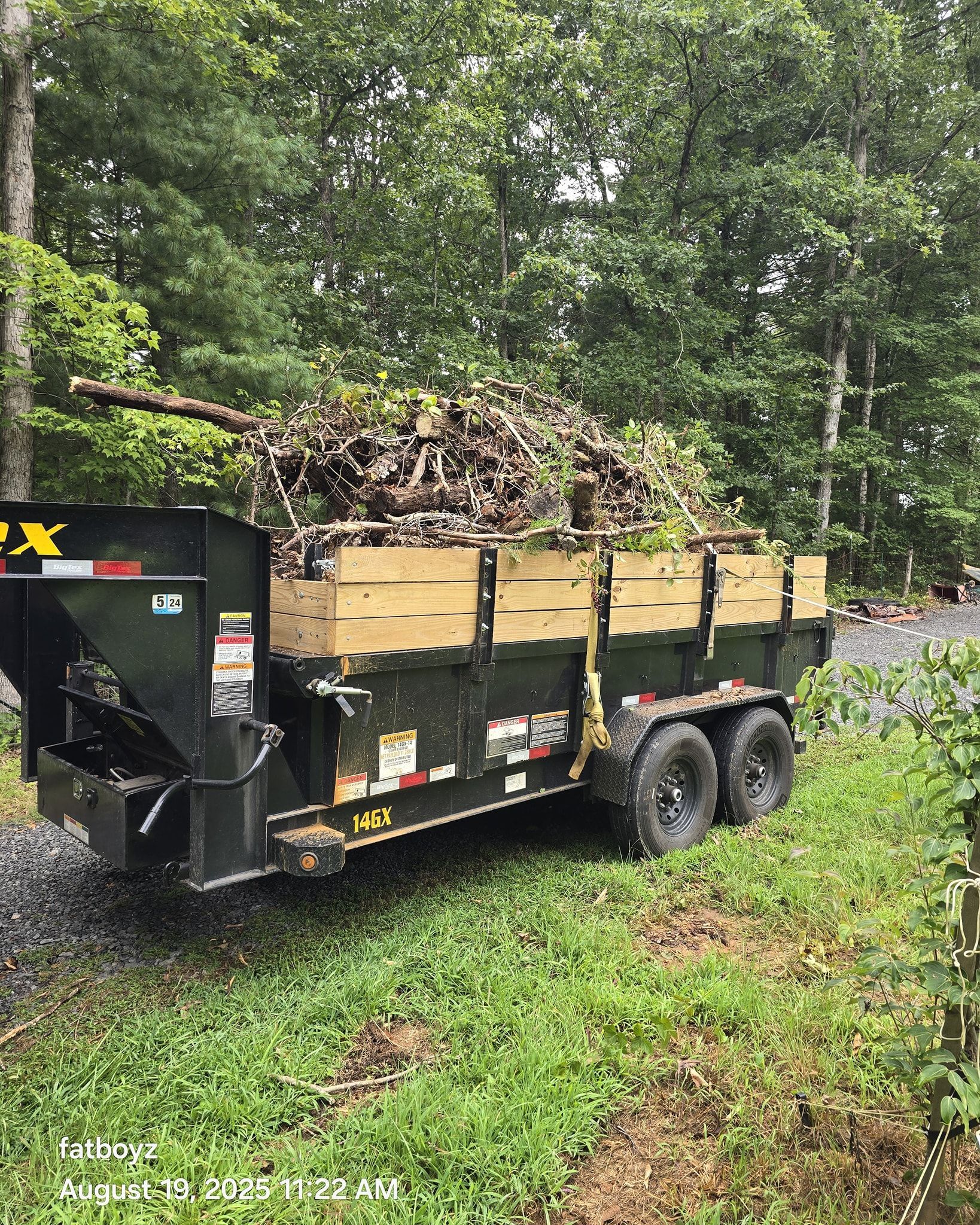 Black trailer loaded with chopped wood on a grassy wooded roadside trail