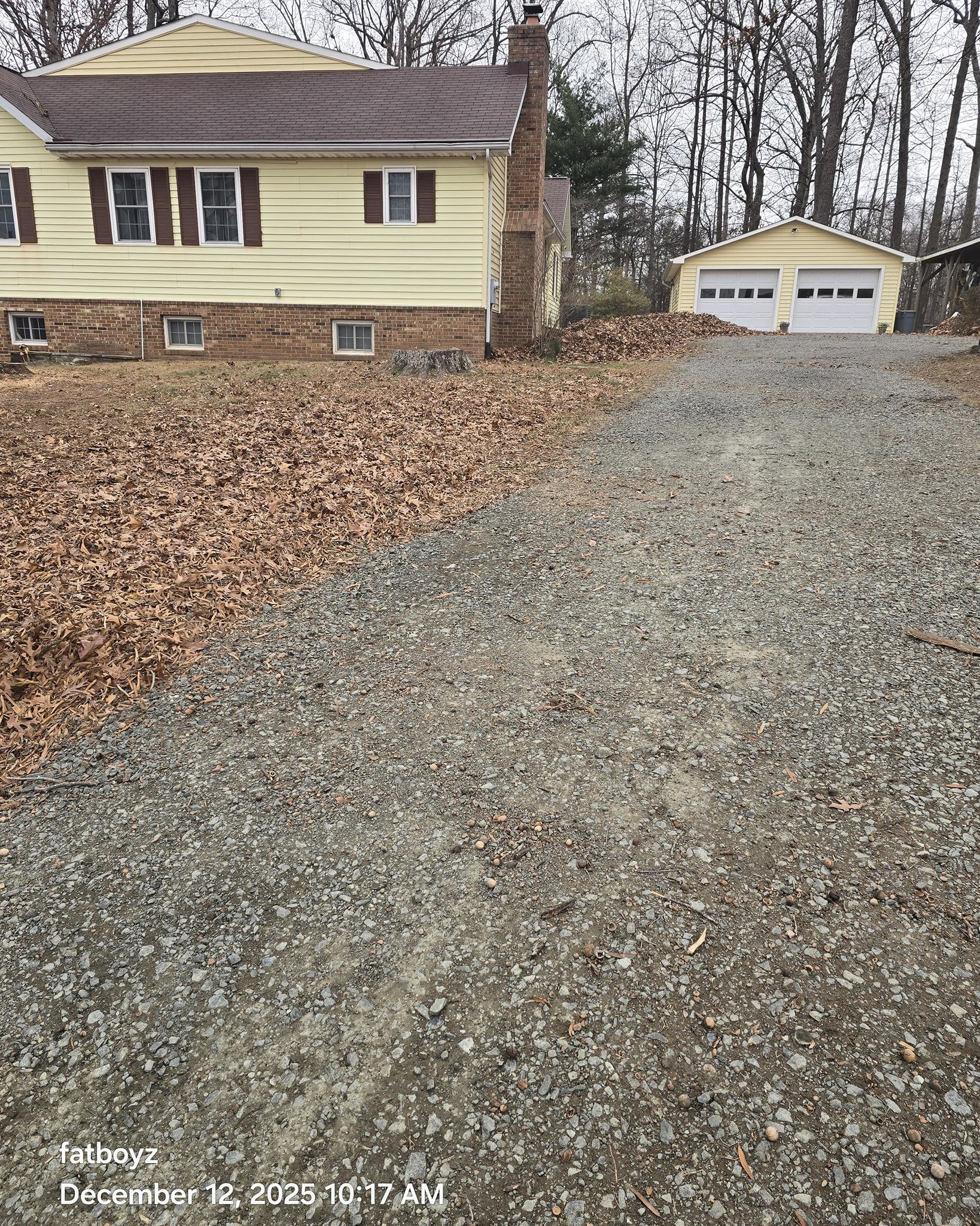 Leaf-covered gravel driveway leading to a beige house and detached garage in a wooded yard