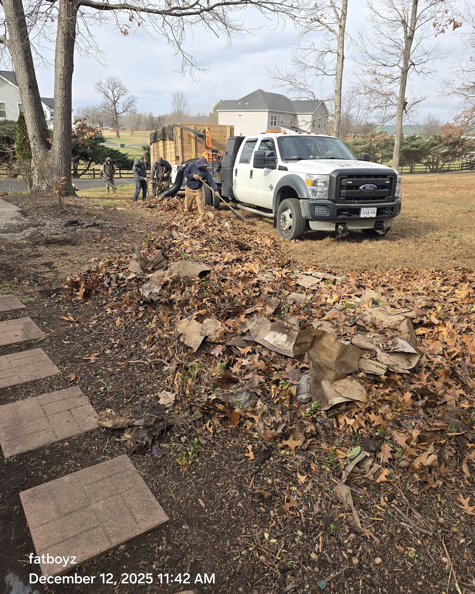White utility truck on a leaf-covered roadside while workers clear debris near a house in autumn.