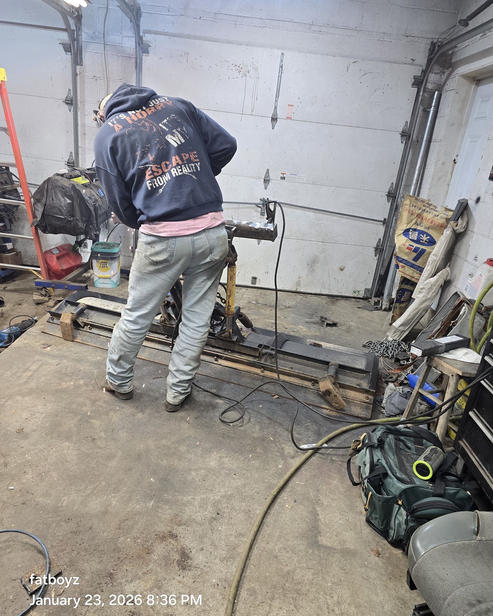 Person in a dark hoodie and gray pants working in a cluttered garage with tools and equipment.