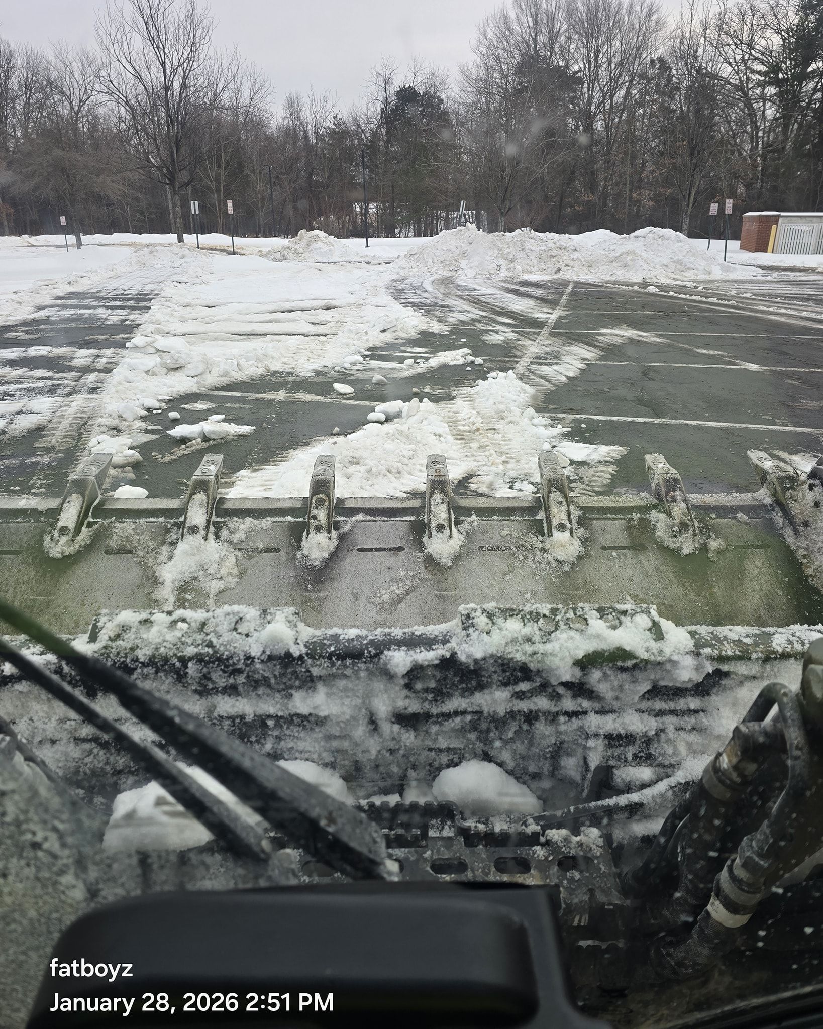 Snow-covered road seen from inside a vehicle, with windshield wipers and trees in the distance.