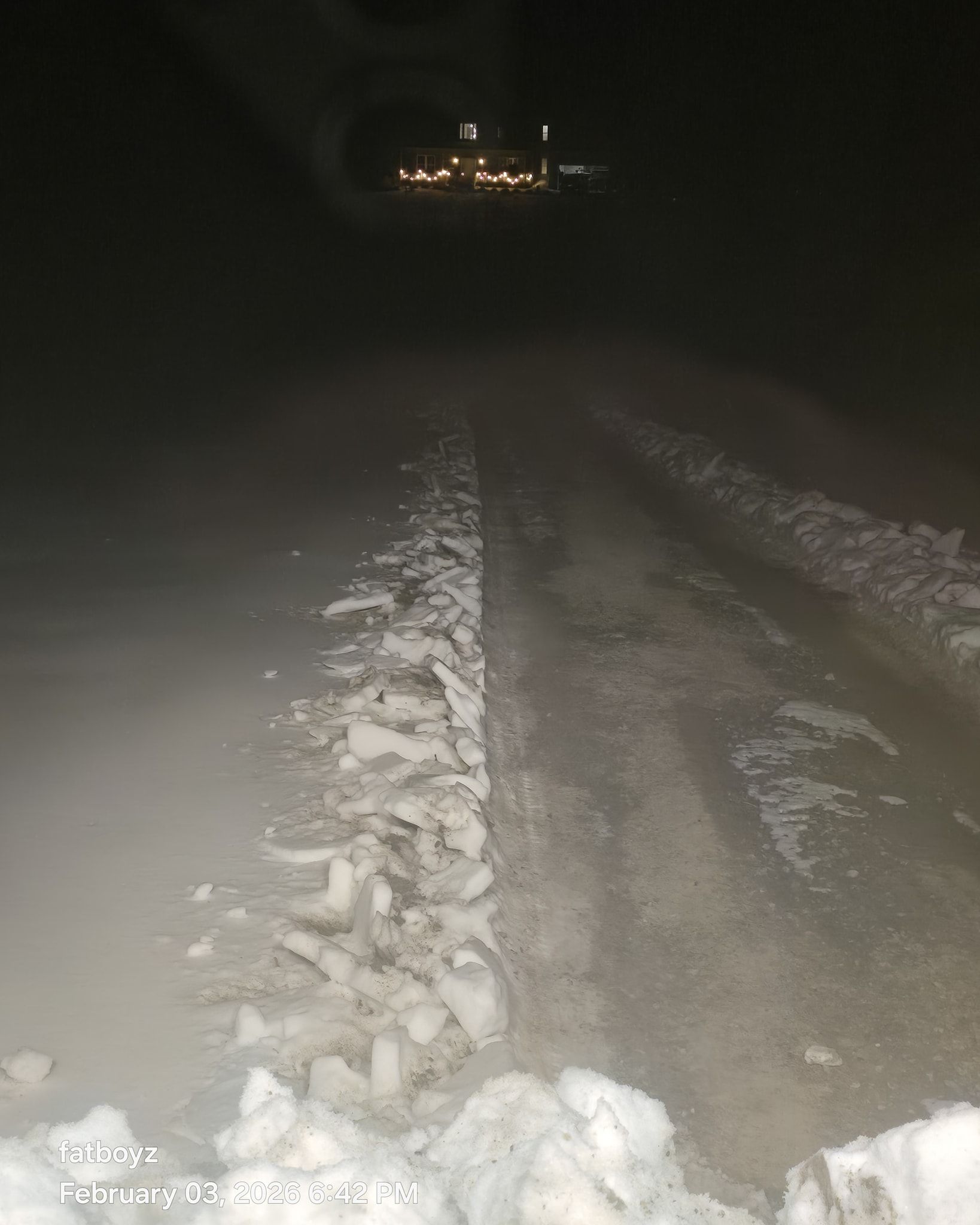 Dark snowy road at night with tire tracks leading toward distant lights.