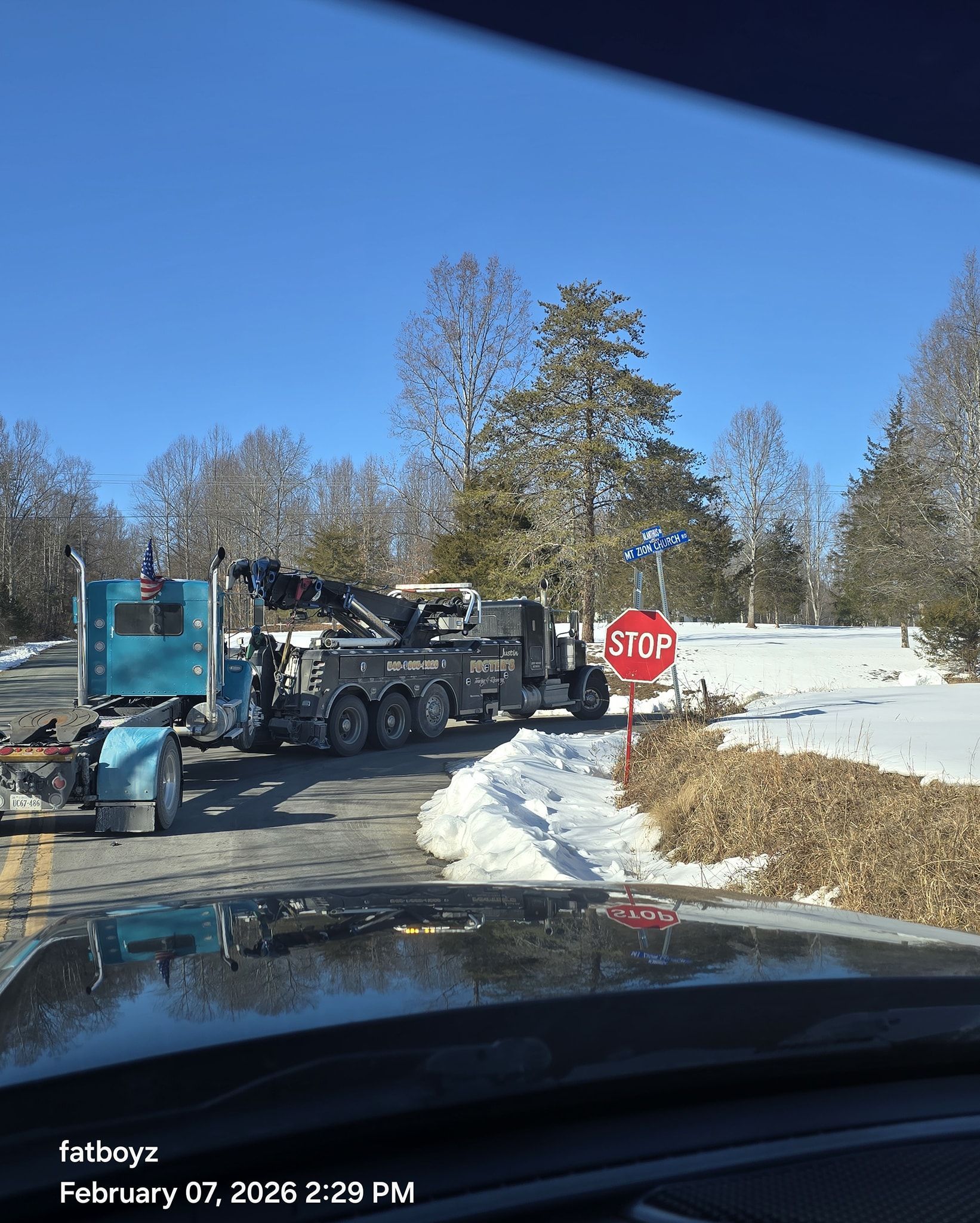 Blue dump truck hauling material across a snowy road near a stop sign, viewed from a car.