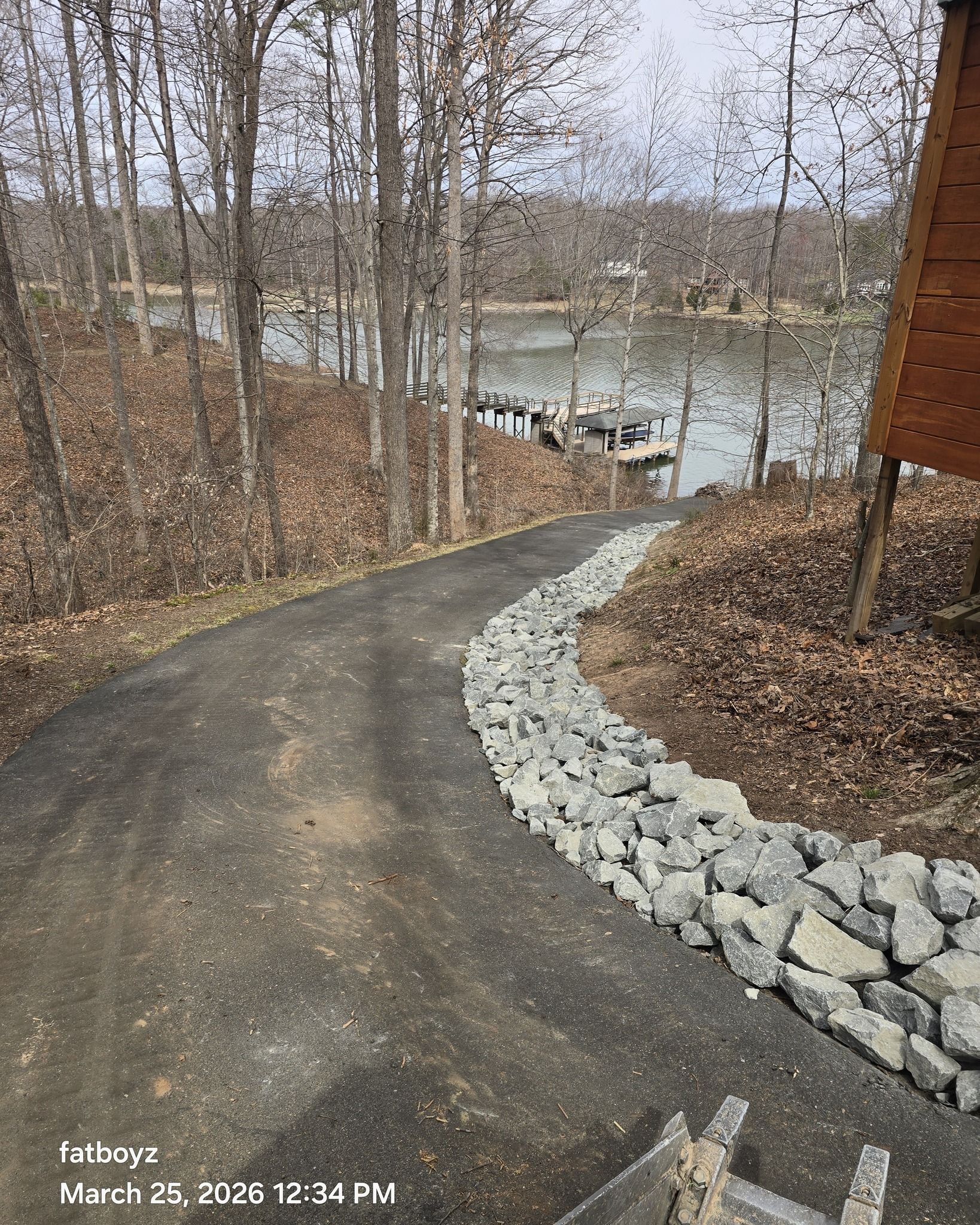 Curving paved path through bare trees leading to a lake, with a rocky border on the right