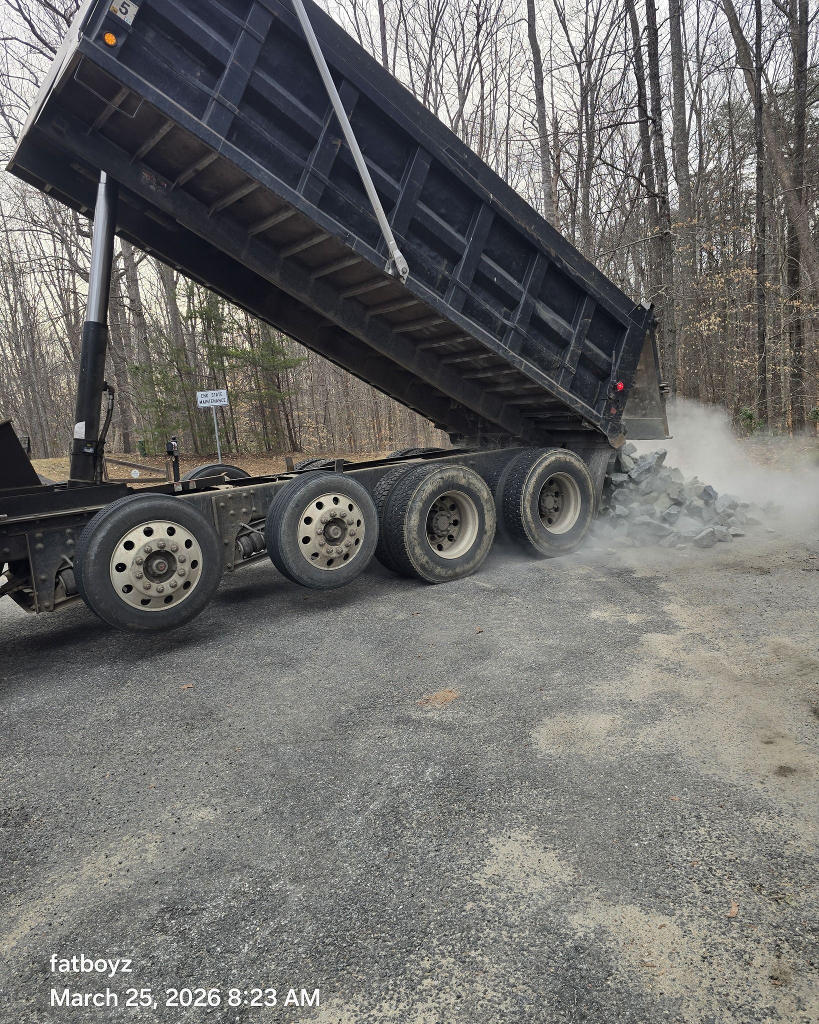 Dump truck bed raised on a gravel road, with dust rising beside a wooded area.