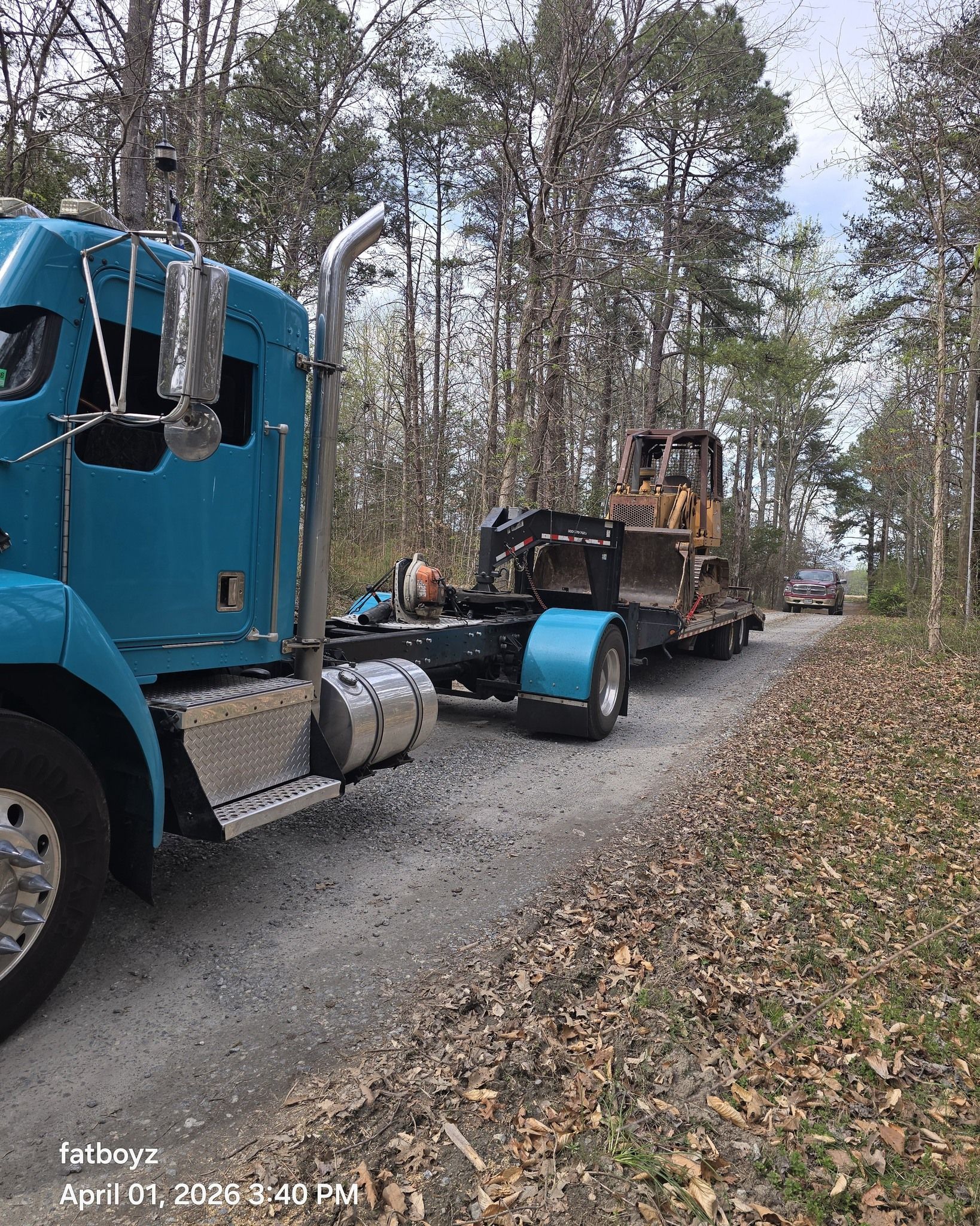 Blue truck hauling a flatbed with a tractor on a gravel road beside a wooded area