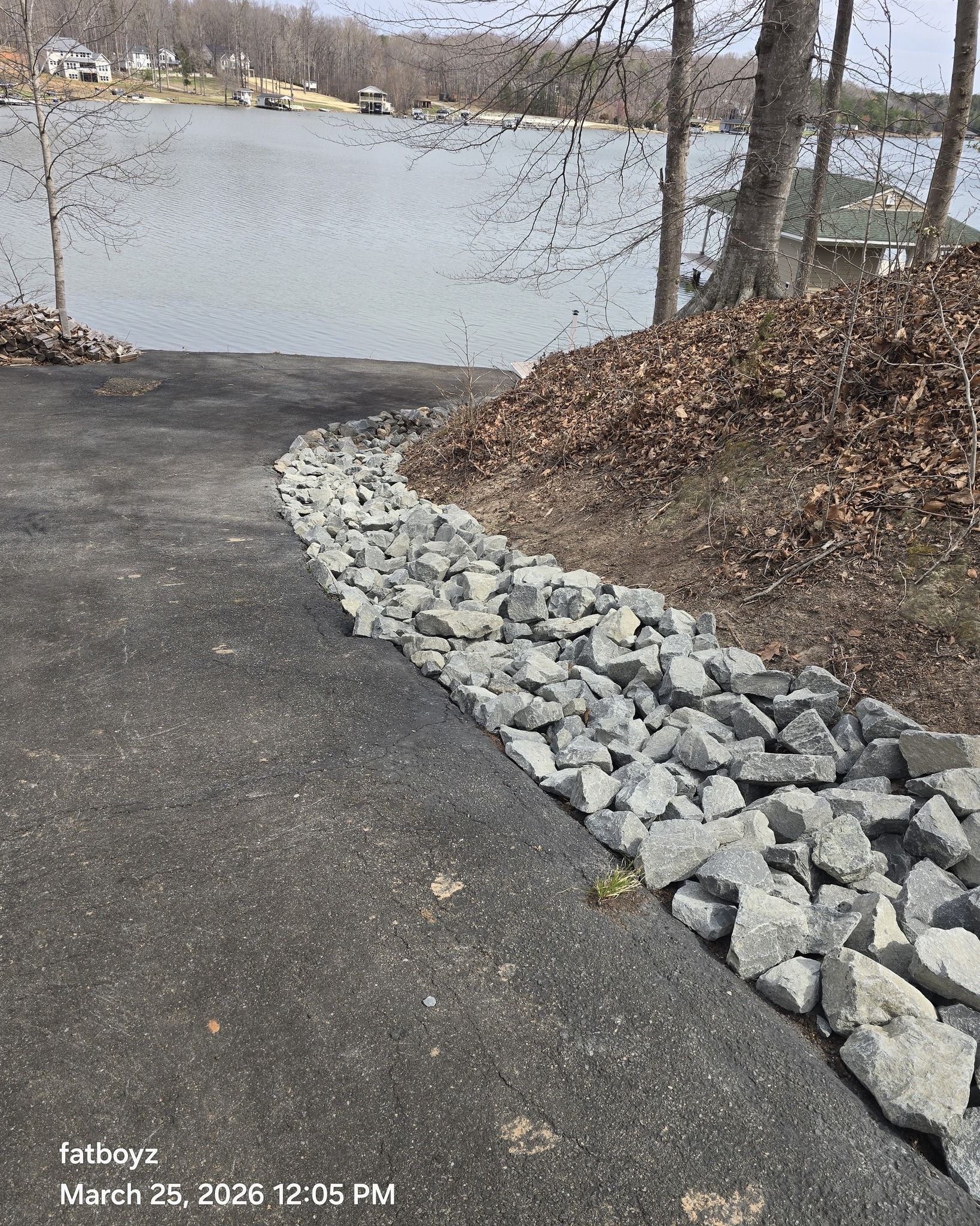 Paved path curving beside a rocky shoreline with a lake and leafless trees in the background
