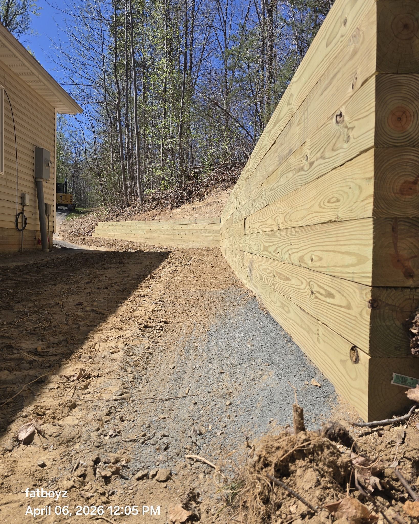 Narrow dirt path beside wood-sided buildings, with evergreen trees and a blue sky in the background.
