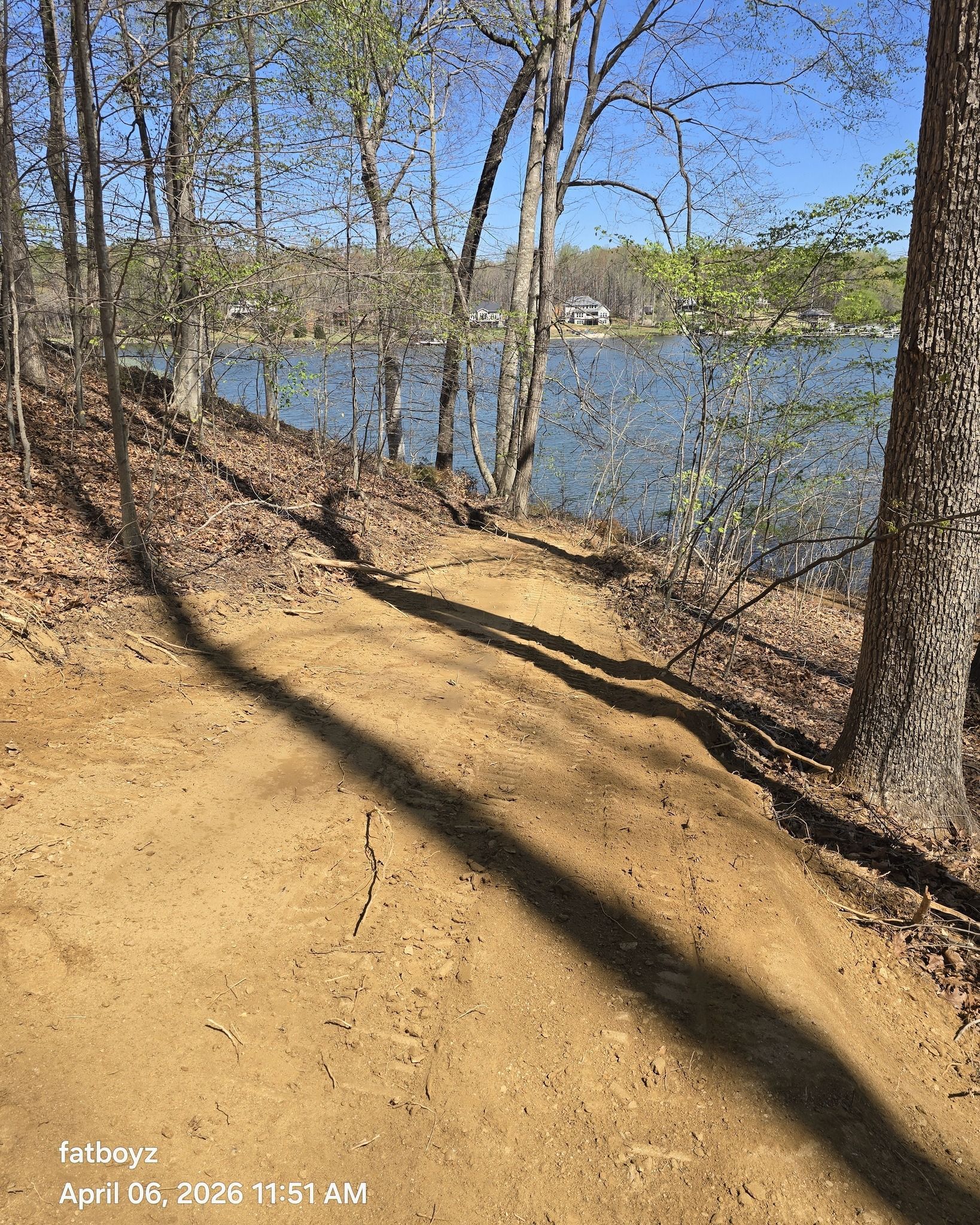 Wooded lakeside trail with leafless trees, long shadows, and blue water under a clear sky.