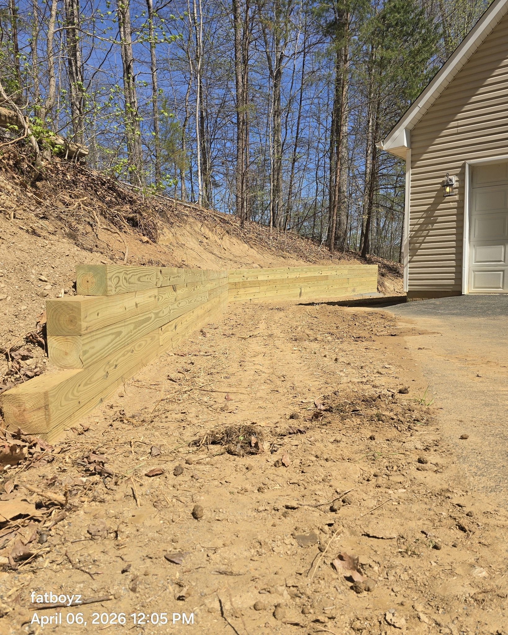 Dirt driveway beside a beige house, with a wooded hillside and tall trees in the background.