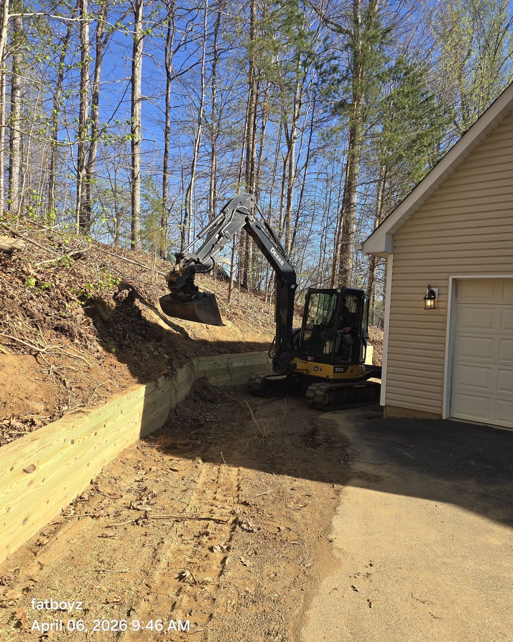 Construction equipment on a dirt driveway beside a house, with a wooded hillside in the background