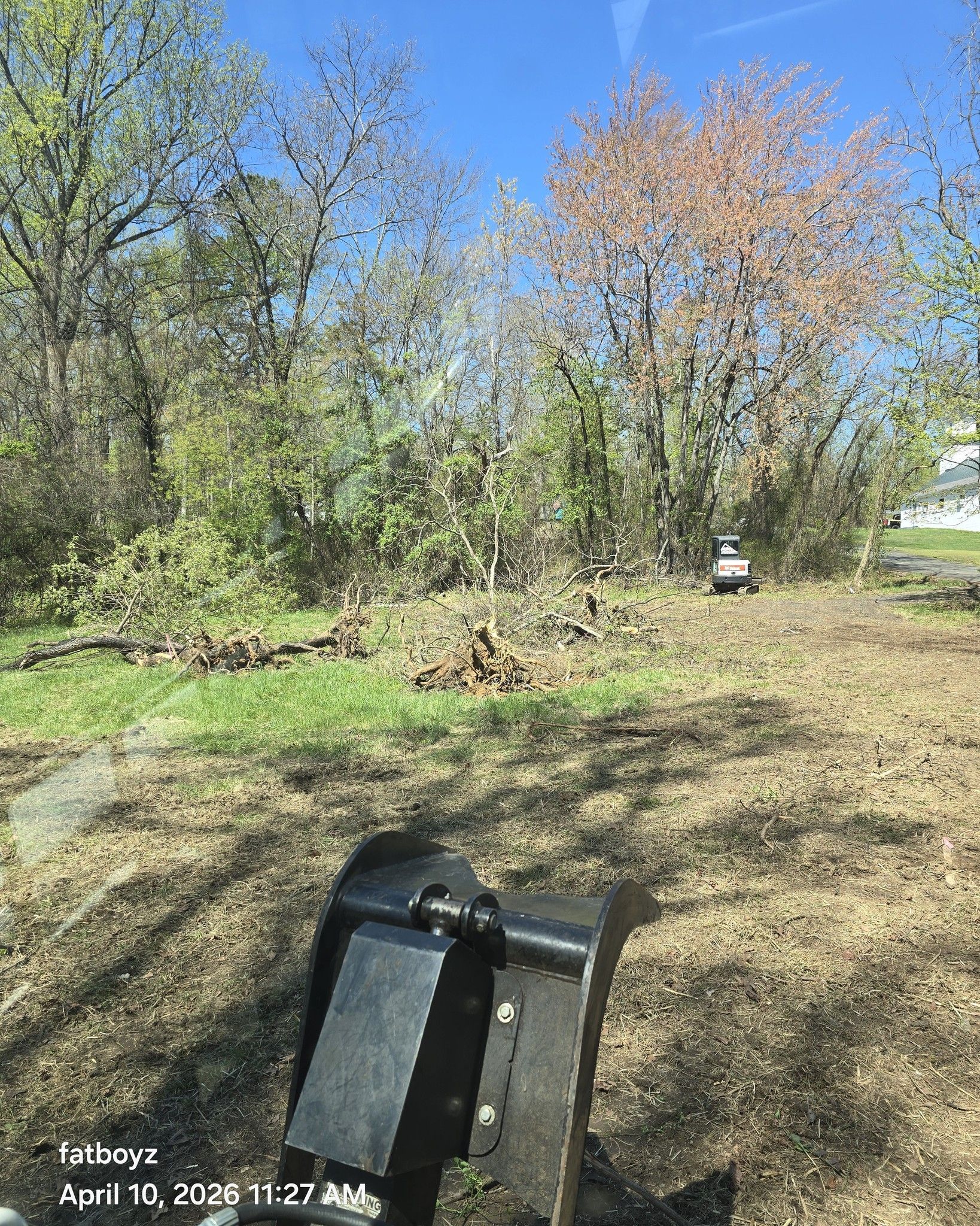 Muddy wooded yard with a black tractor bucket in the foreground under a blue sky