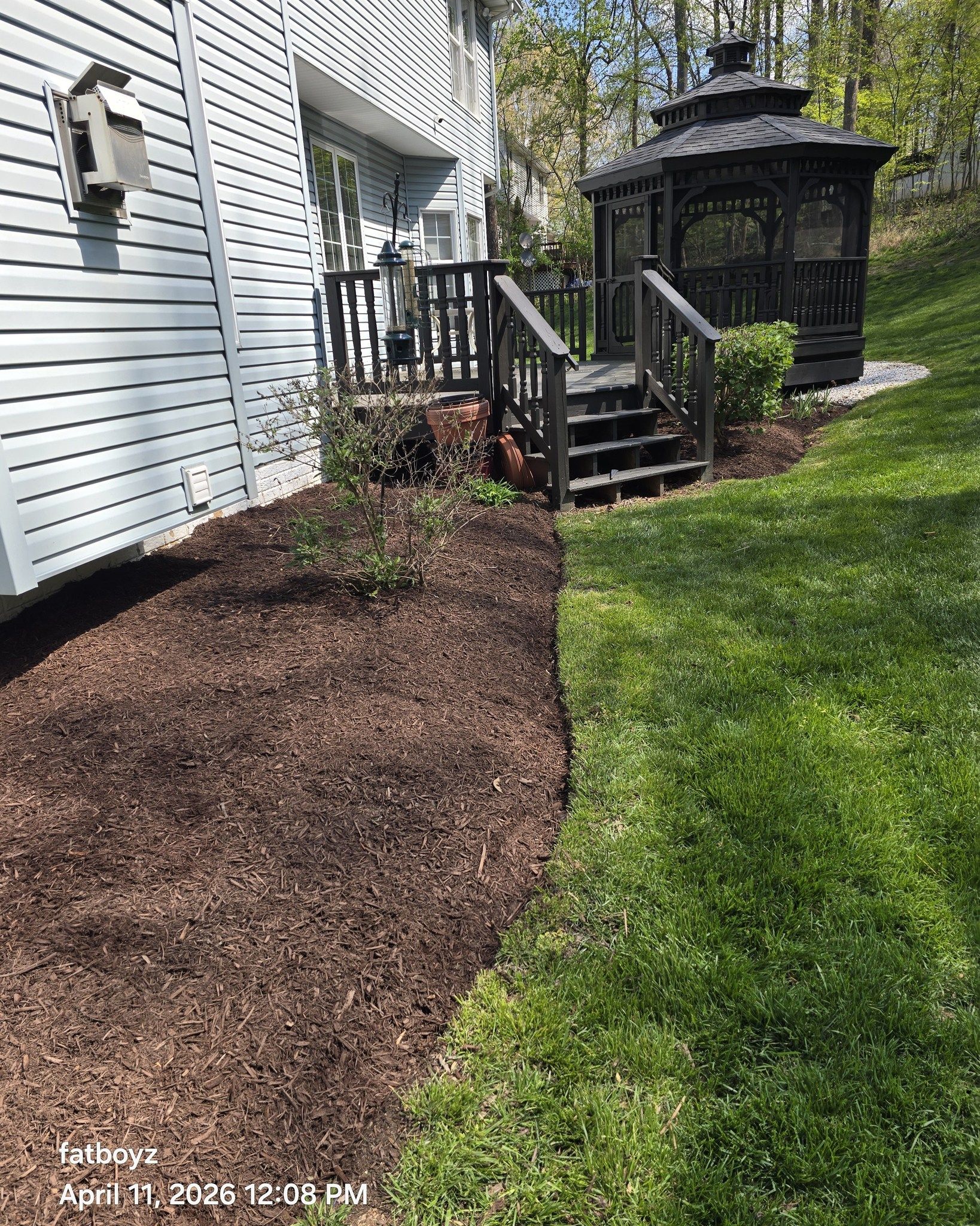 Mulched garden bed along a white house beside a green lawn and small black gazebo.