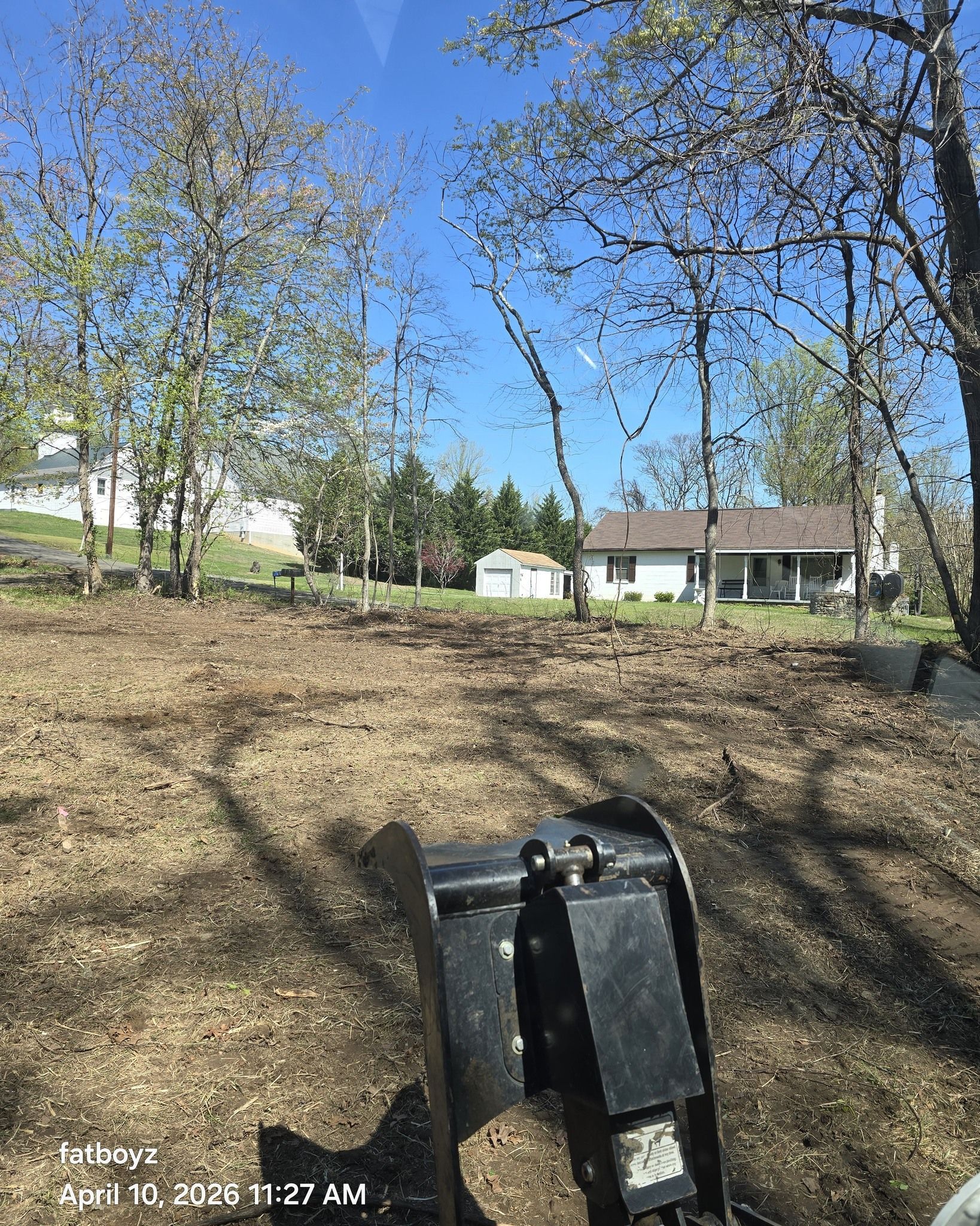 Sunny backyard with bare trees, a house in the distance, and a black trash bin in the foreground.