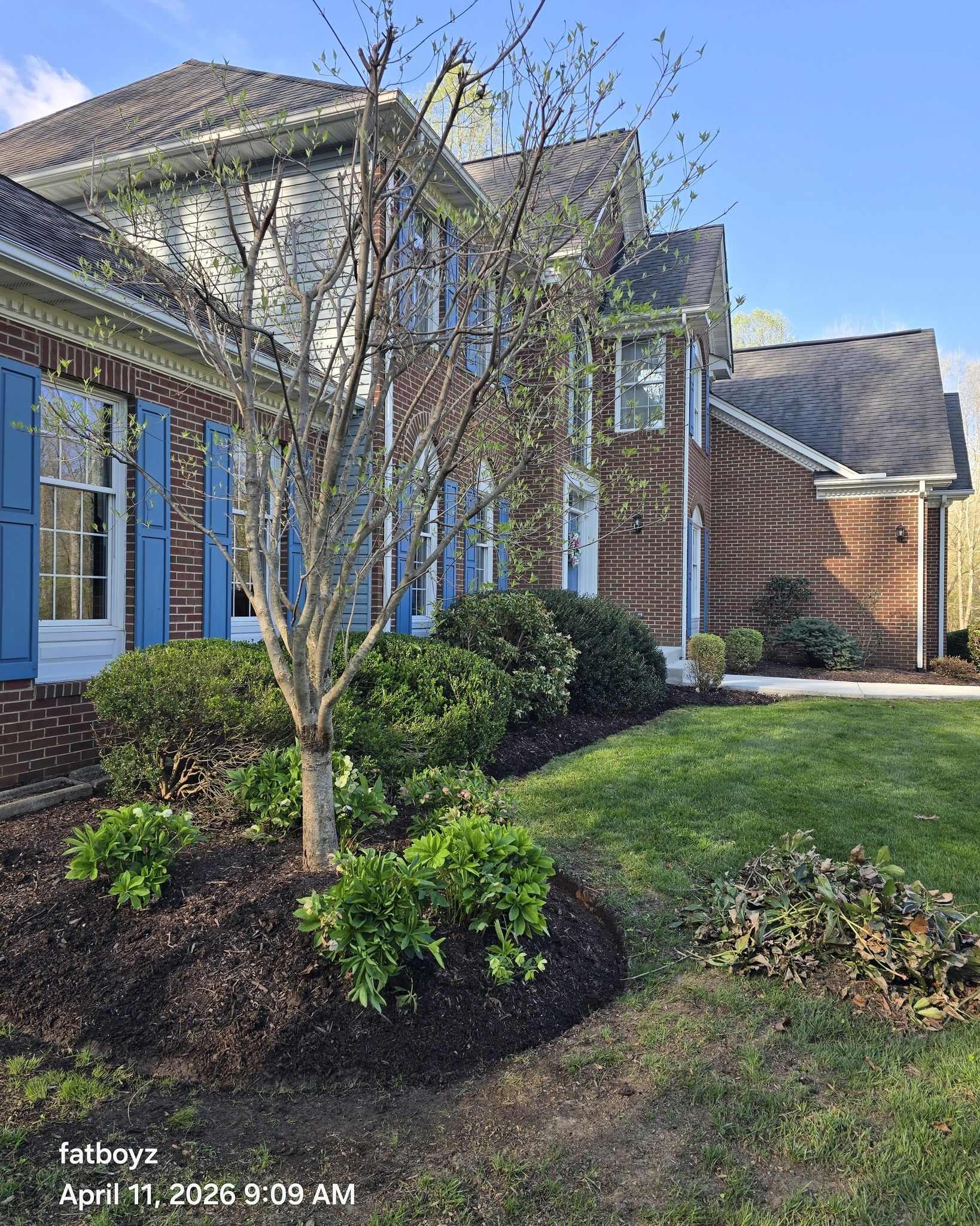 Suburban house exterior with brick and siding, blue shutters, landscaped shrubs, and a front lawn