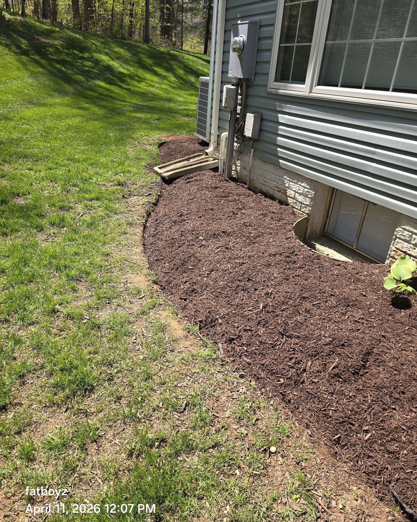 Mulched landscaping bed along the side of a house beside green grass and utility meters