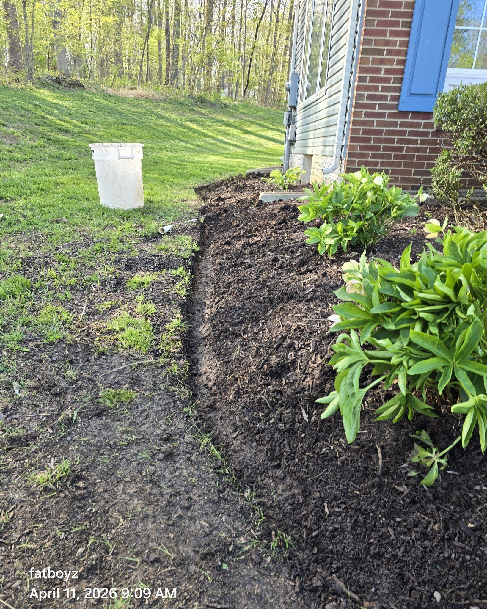 Freshly dug mulch bed curving beside a house, with shrubs and a white bucket on a grassy slope.