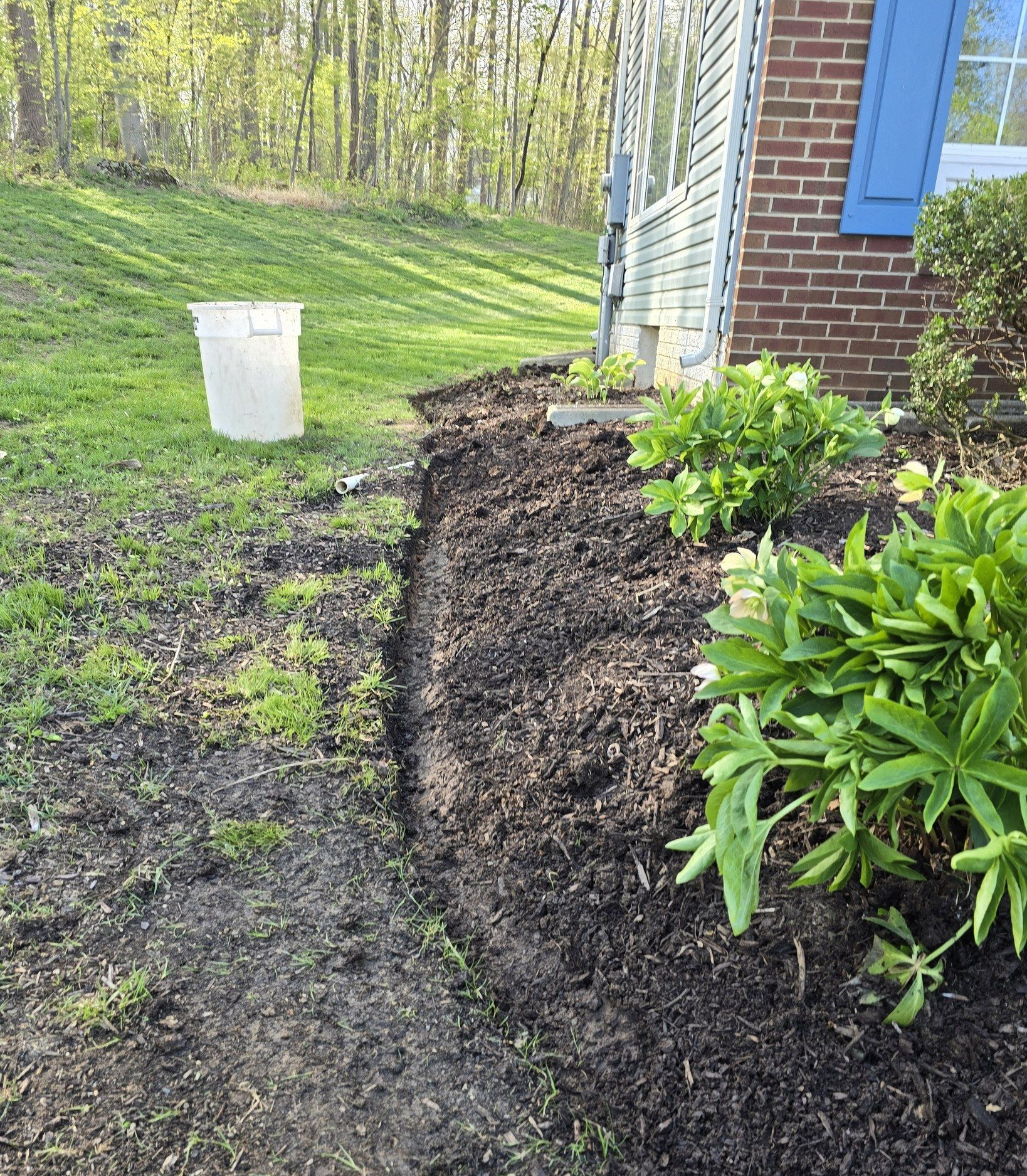 Freshly mulched garden bed beside a brick house, with green shrubs and a white bucket on a grassy slope.