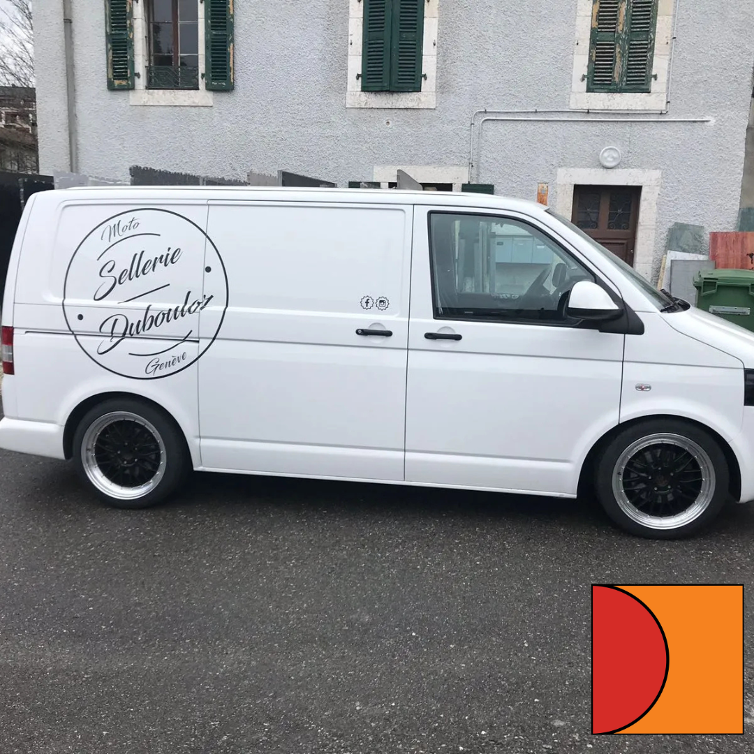 Une camionnette de livraison blanche est garée dans une rue, à côté d'un bâtiment en pierre aux volets verts.