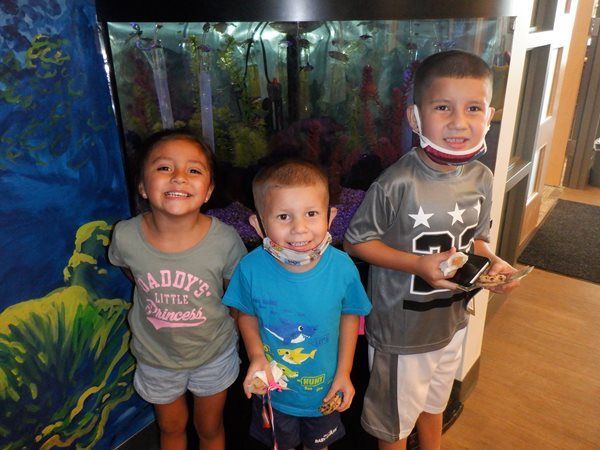 Three smiling children stand in front of a fish tank. 