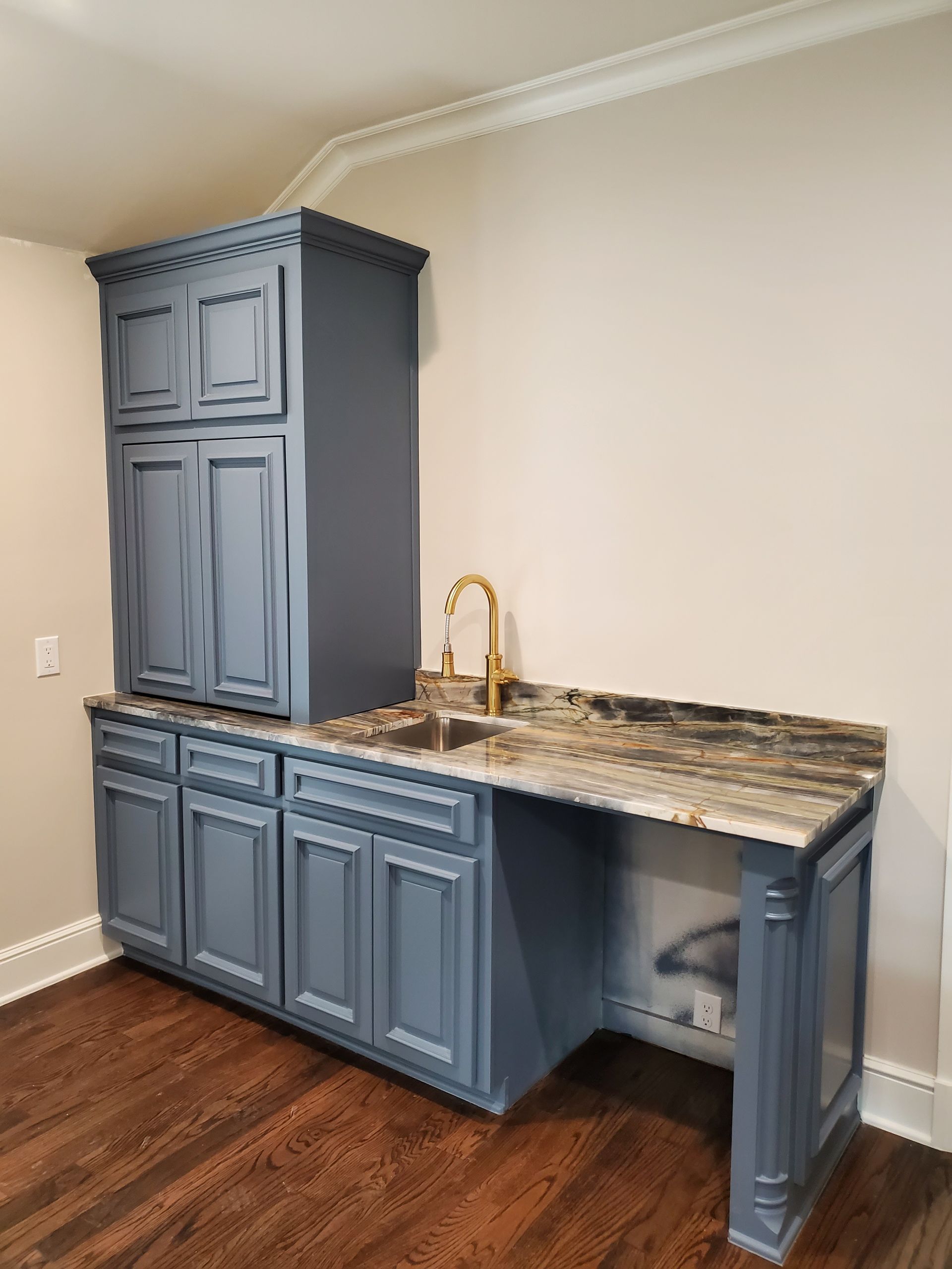 Blue cabinetry with a sink and countertop. Gold faucet. Dark wood floor and cream-colored walls.