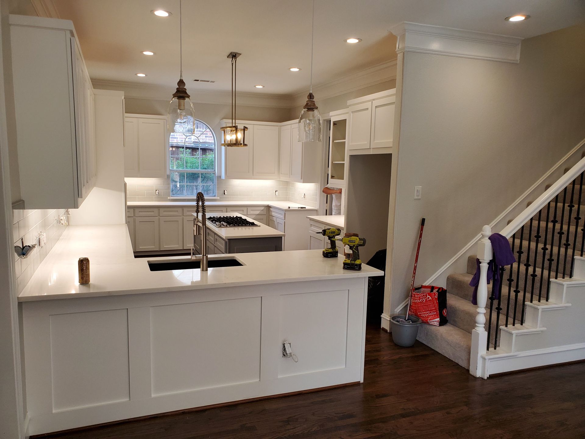 Modern white kitchen with island, cabinets, countertops, and pendant lights; staircase visible.