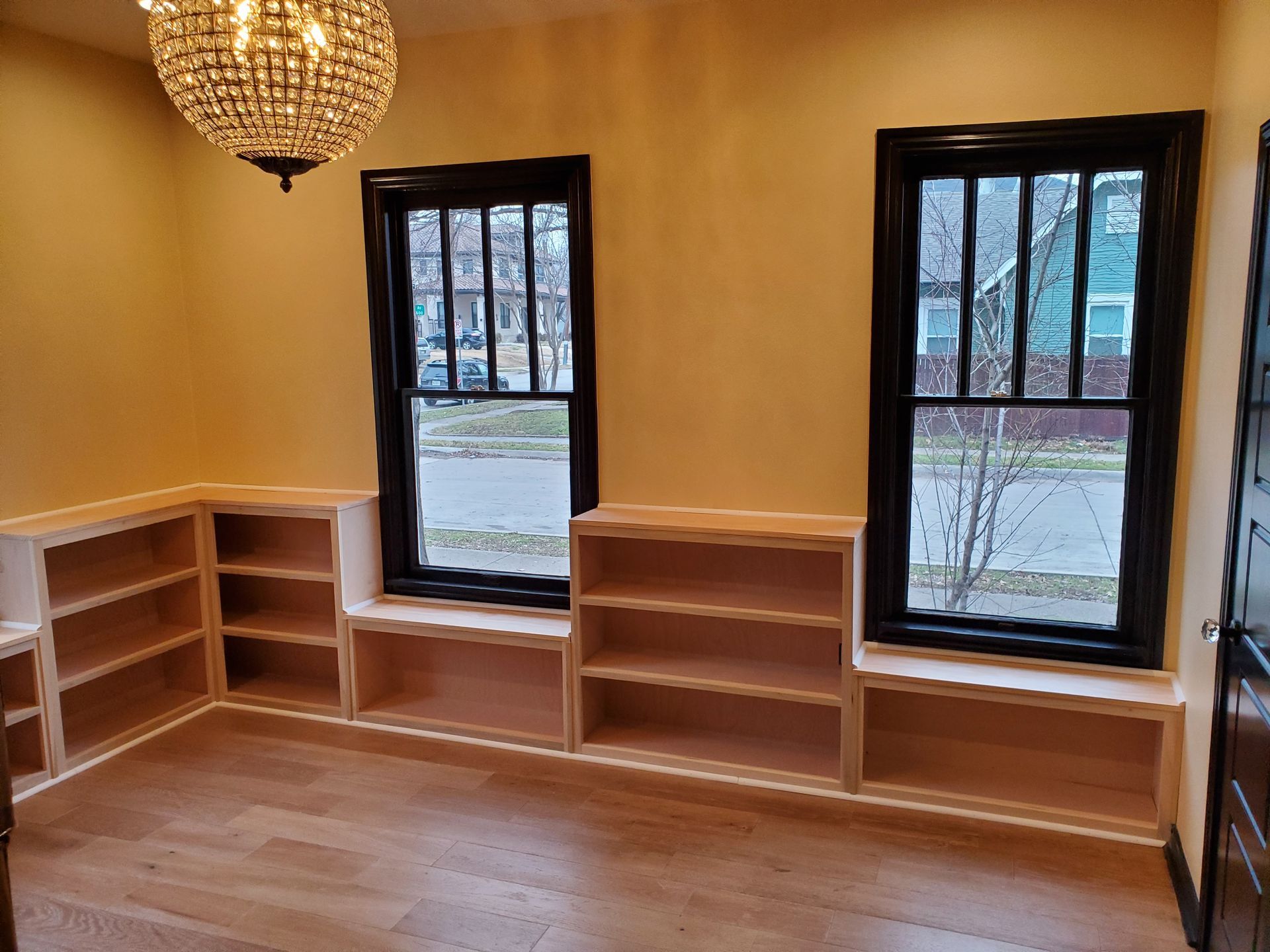 Interior room with two windows, bookshelves, hardwood floor, and a chandelier. Walls are yellow, windows have black frames.